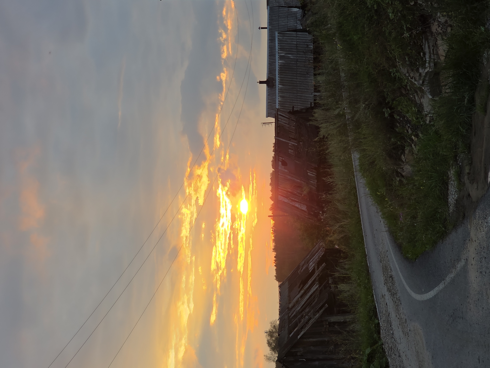 The image depicts a rural sunset scene with a winding dirt road in the foreground. To the left of the road are two dilapidated wooden structures, one appearing to be a shed or outbuilding with a collapsed roof. To the right, a row of corrugated metal-roofed structures, possibly houses or barns, are visible. The sky is filled with dramatic clouds illuminated by a bright orange sunset. Power lines stretch across the sky. The overall impression is one of a somewhat neglected but picturesque rural setting. This scene is located in the city of Magnitka, Russia.