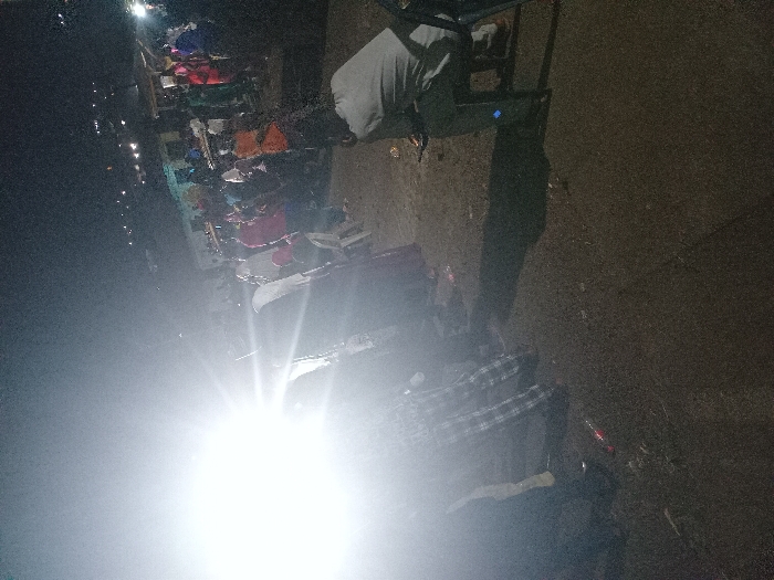 A dark and blurry scene shows a street market at night. Stalls are lined with colorful textiles or clothing. In the foreground, a bright light obscures details, but a person in a plaid shirt and dark pants is partially visible. Behind the individual, the ground appears to be rough, possibly dirt or gravel. A person stands near a cart or table structure, their back facing the viewer. The overall impression is one of an active, though poorly lit, marketplace.
