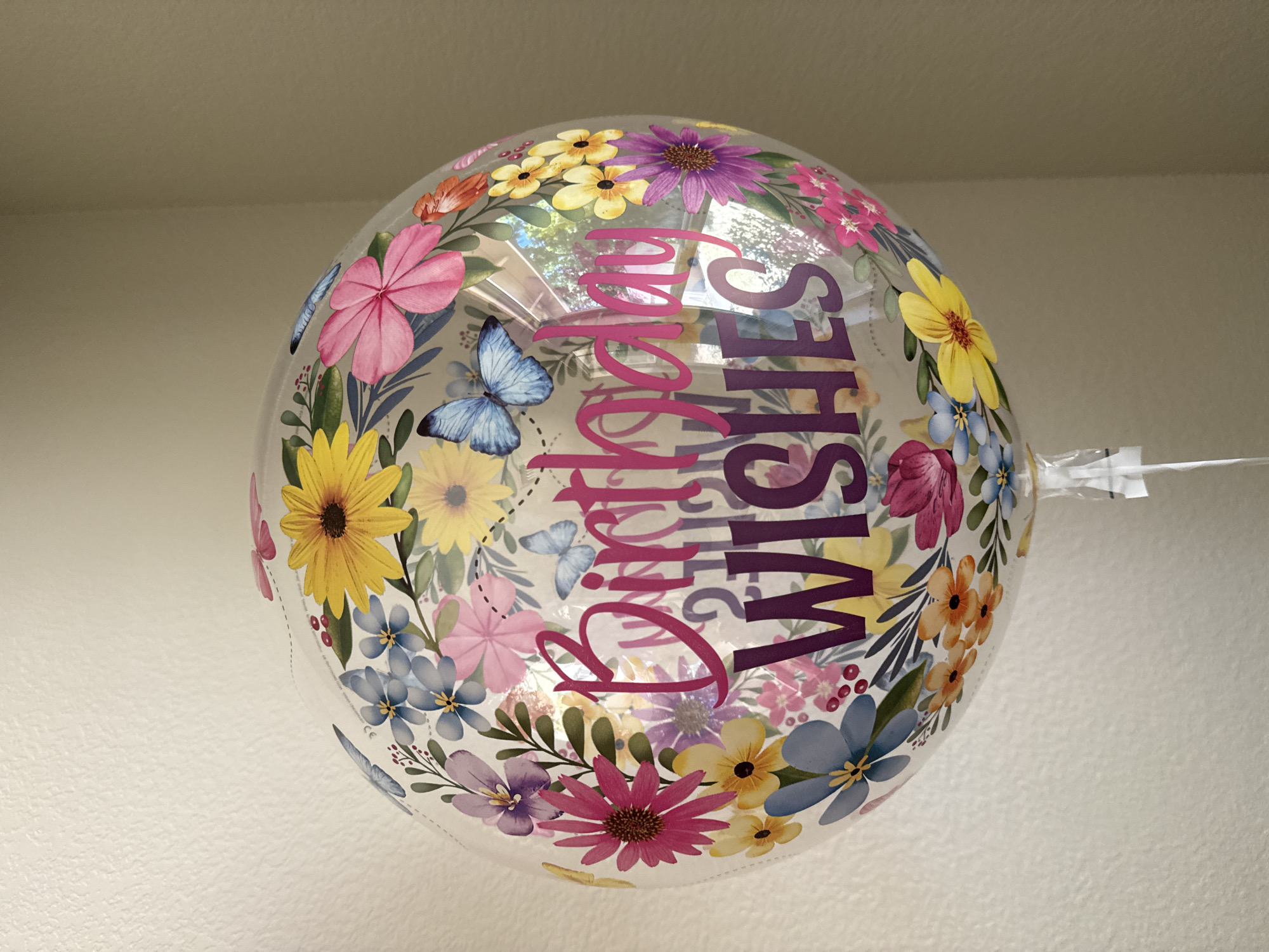 A clear, spherical balloon with a floral and butterfly design is the central subject of this image. The balloon is decorated with various colorful flowers, including pink, yellow, purple, and blue blossoms, as well as blue butterflies. The words "Birthday Wishes" are printed in large, pink and purple letters across the front of the balloon. The balloon is suspended by a clear plastic ribbon, which extends downwards out of the frame. The background consists of a textured, light-colored wall, possibly painted. The lighting suggests an indoor environment with ambient light. The image was taken in West Sacramento, United States of America.