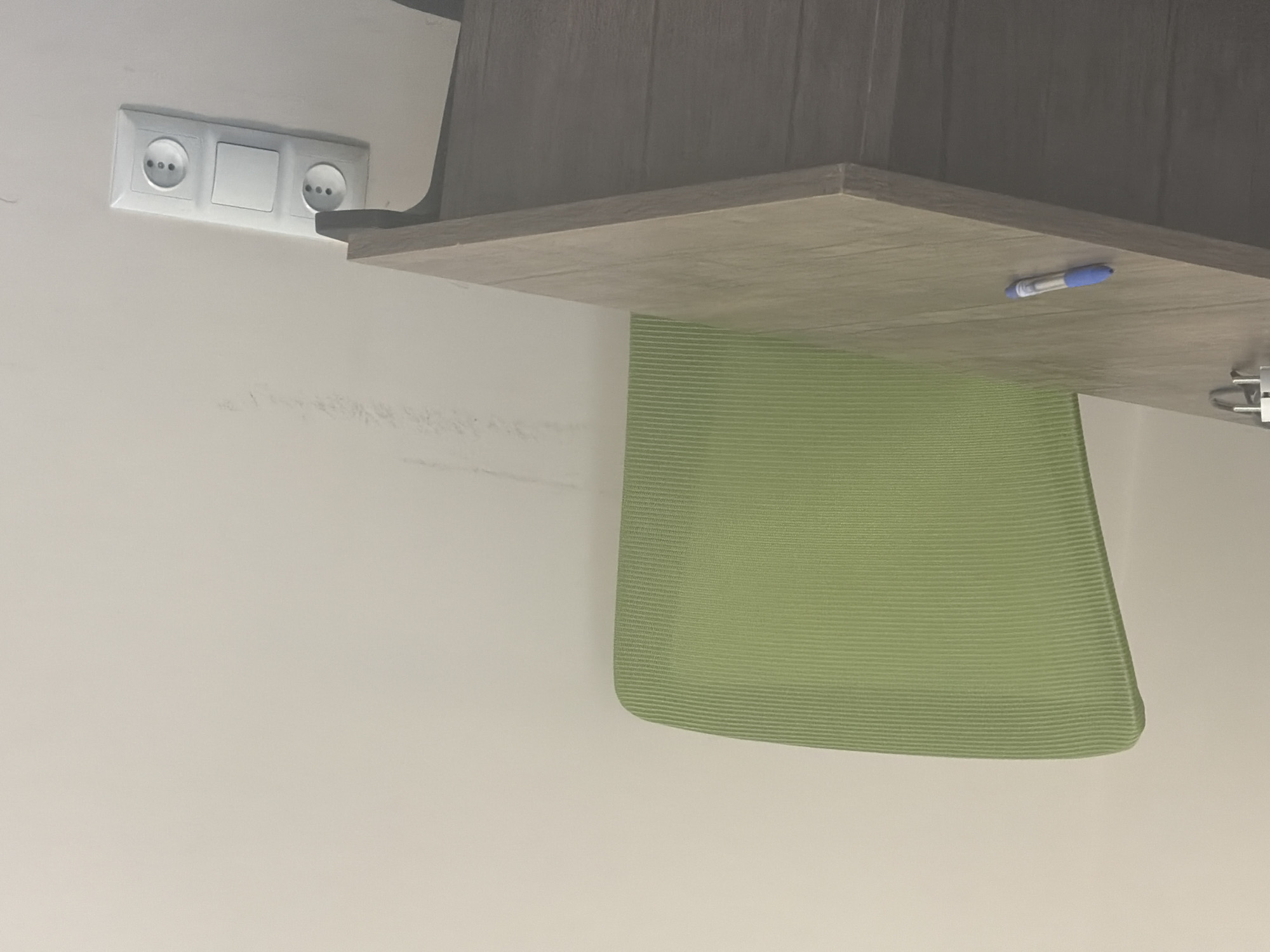 The image shows a wall-mounted electrical outlet with two sockets and a switch, positioned to the left of a wooden desk. Below the desk, a light green mesh chair is visible, partially obscuring the wall. A blue pen rests on the surface of the desk, to the right of the outlet and near the edge of the desk. The scene appears to be indoors, likely in an office or a home setting, with neutral-colored walls. The lighting suggests an indoor environment, and there are no indicators of the time of day or weather. The image is oriented vertically, with the desk and chair appearing at the top and bottom respectively, implying the photograph was taken with the camera rotated. No text is visible in the image. This scene is consistent with an urban setting like Yerevan, Armenia, where such electrical fittings and office furniture are common.