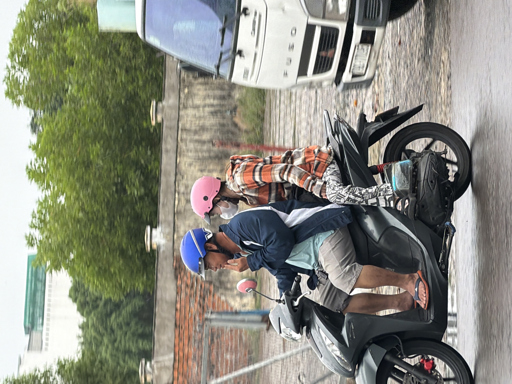A side view of two people on a scooter moving on a wet road in Nha Trang, Vietnam. The person in front, a male, wears a blue helmet, a dark blue and white jacket, and light-colored shorts. He is touching his chin with his right hand. The person behind, a female, wears a pink helmet, an orange and white plaid jacket, and patterned leggings. She wears a white face mask. A white truck with the brand name "FUSO" visible on the front is to the right of the scooter, in motion. The background shows a brick wall, green trees, and a building. The road surface is wet, indicating recent rain or mist.