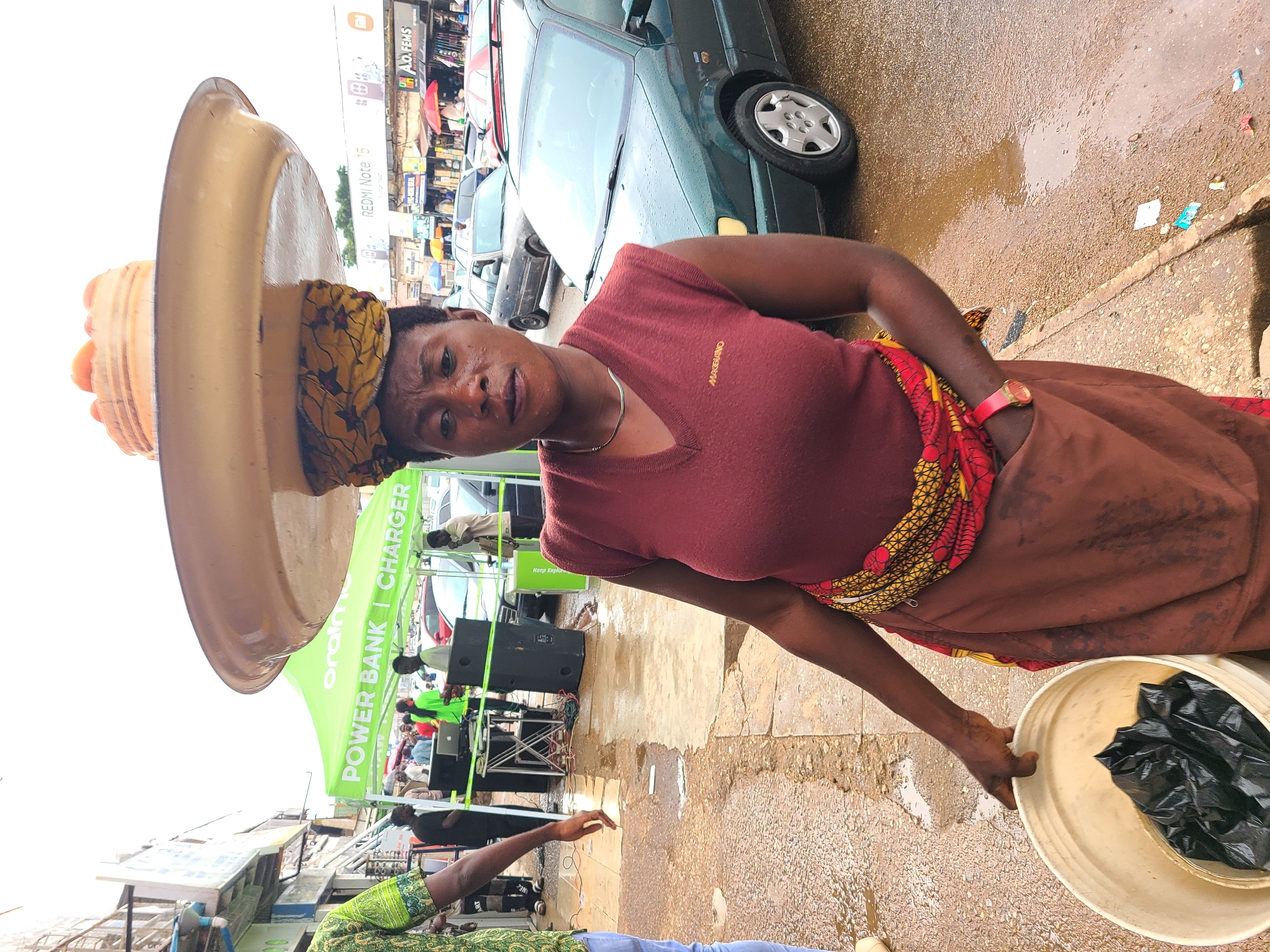 A woman in Jos, Nigeria, balances a large tray of fruit on her head. She wears a maroon sleeveless top over a patterned skirt and a brown skirt tied around her waist. She holds a white bucket containing black plastic bags in her left hand. The scene is outdoors, with a wet paved ground and several vehicles in the background. A green tent with "ORAIM POWER BANK | CHARGER" written on it is visible. Other people and stalls are present in the distance, suggesting a market or busy street. The sky is overcast.