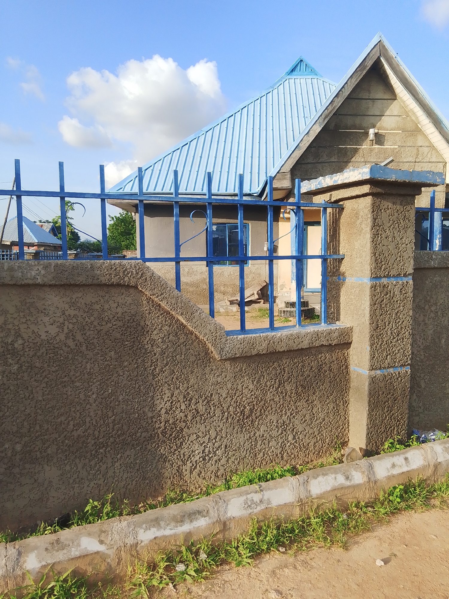 This image depicts a close-up view of a textured concrete wall and a blue metal fence in front of a building with a blue corrugated metal roof. The building appears to be a house, with visible windows and a doorway. The scene is set outdoors, with a clear blue sky and some white clouds visible in the background. Green grass is growing along the base of the wall and fence. The time of day appears to be late afternoon or early evening, given the angle of the sunlight casting shadows. The location is Jalingo, Nigeria. There is no visible text in the image. The overall mood is peaceful and serene.