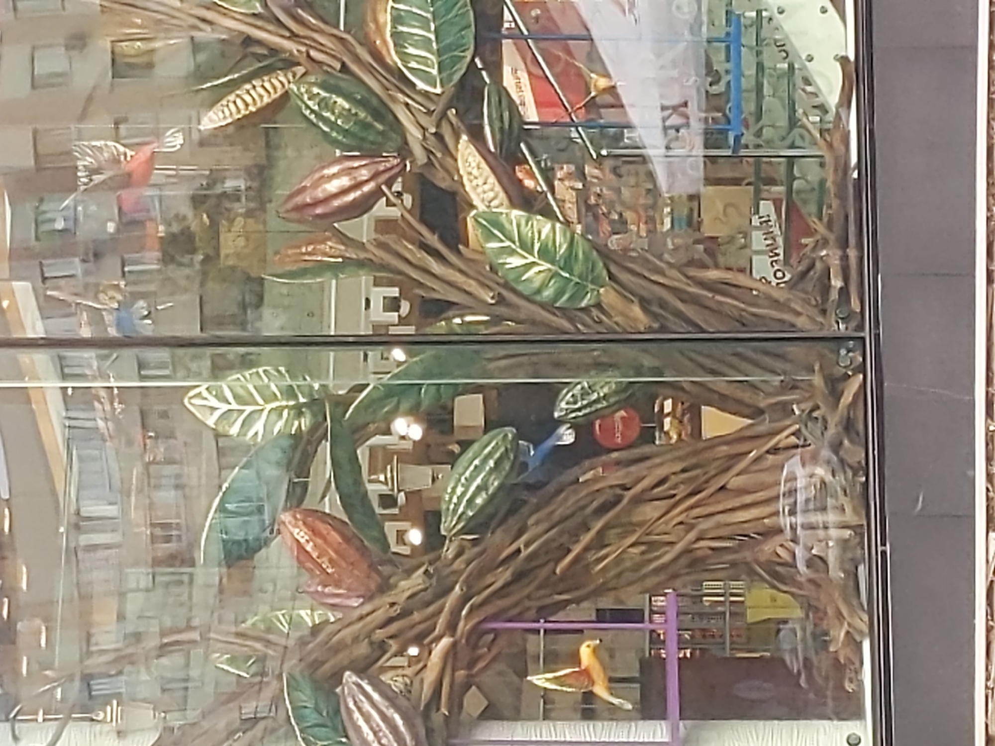 The image shows a storefront display featuring a large, artificial tree with branches resembling cacao pods and leaves. Two small, decorative birds are placed on the display, one yellow and one blue. The background of the display reflects the exterior of buildings, suggesting it is located in an urban environment. The presence of a yellow Ukrainian flag banner on the right side of the image, along with Cyrillic text on some of the reflections, indicates the location is likely in Kyiv, Ukraine. The storefront itself is made of glass, allowing a view into the display and the reflected surroundings.
