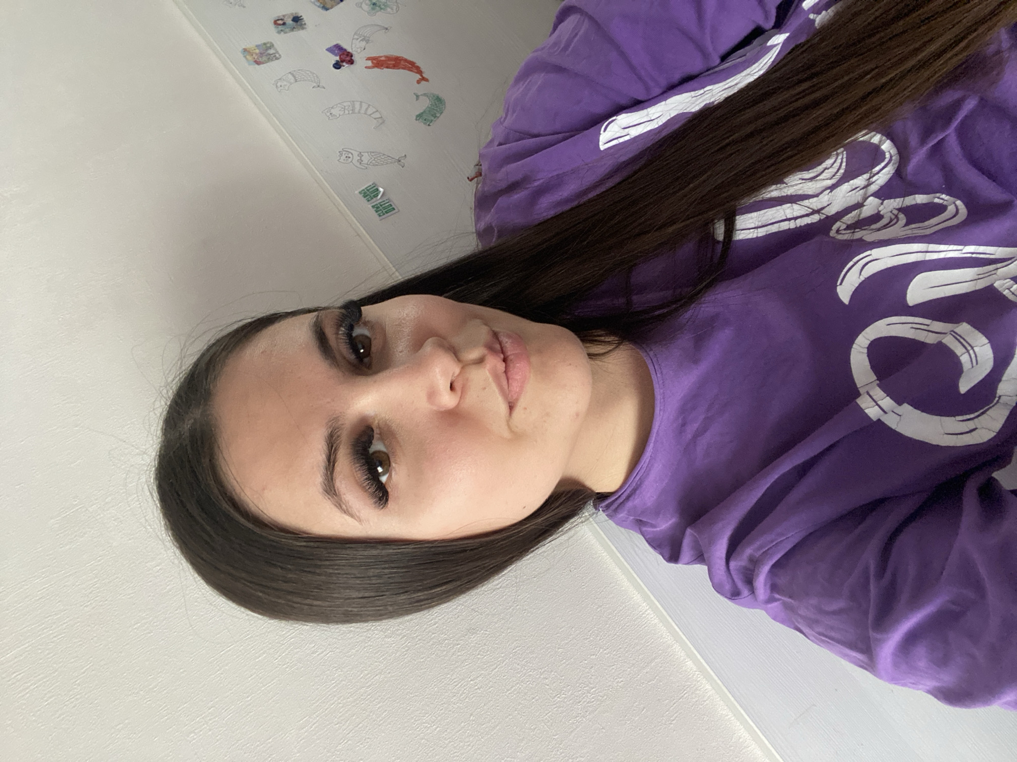 A young woman with dark hair and long eyelashes is wearing a purple long-sleeved shirt with white lettering. She is positioned indoors, with a white wall featuring cartoon-like stickers in the background. The exact location in Chaikovsky, Russia, cannot be determined from the image alone.