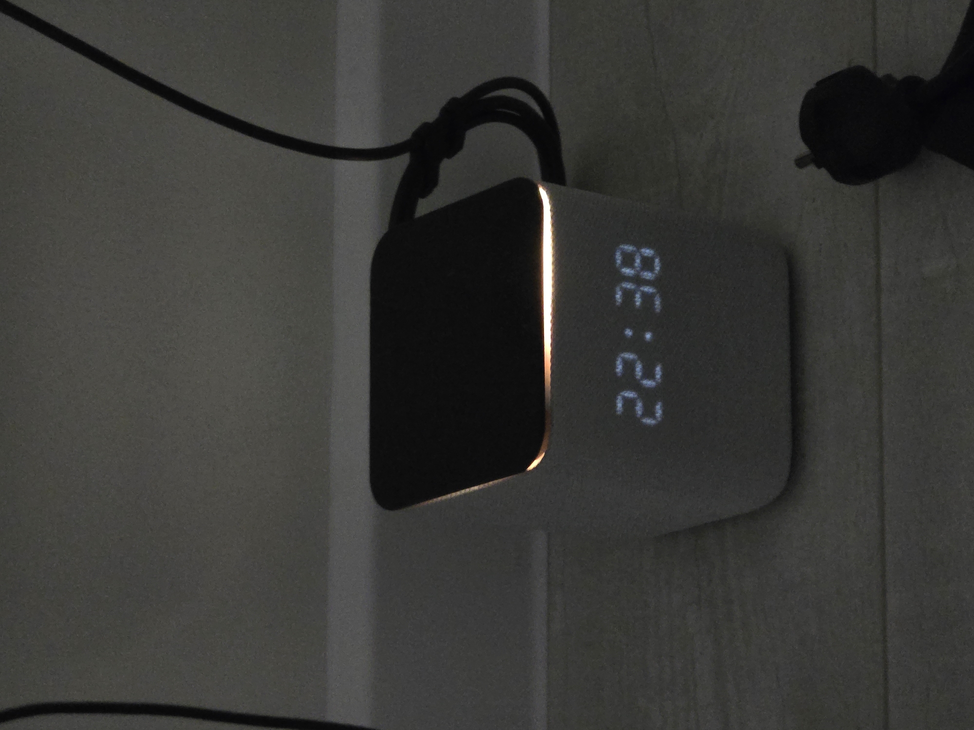 The image depicts a cube-shaped digital alarm clock displaying the time "22:38". The clock is light gray, with a dark screen on one face. There is a black power cord connected to the top of the clock. The clock is mounted on a light-colored vertical surface, potentially a wall. The image is dimly lit. There is no identifiable location in the city of Chaikovsky, Russia discernible from the provided image.