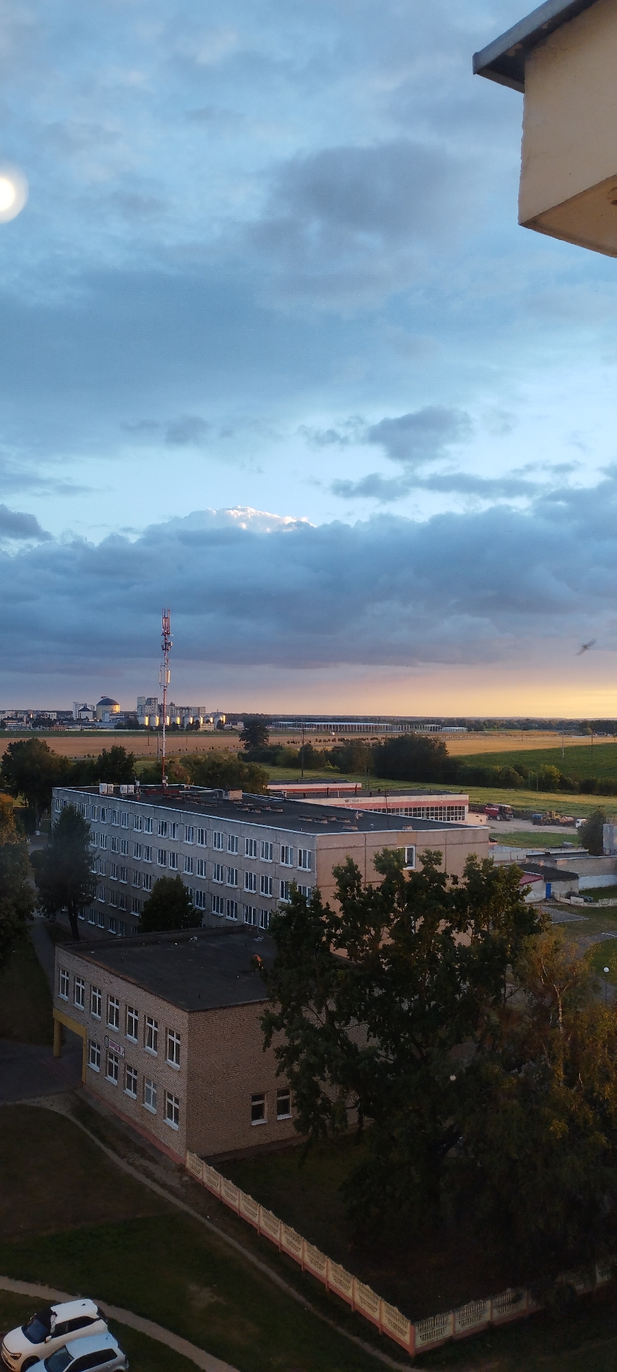The image shows a broad aerial view of a semi-urban area at dusk, likely in Skidzieĺ, Belarus. The sky is a mix of light and dark clouds, with the sun setting on the horizon, casting a warm glow. In the foreground, two buildings are visible: a larger, grey, multi-story building with numerous windows, and a smaller, brick building with a yellow facade on the lower section. Both appear to be institutional or residential. A white fence runs along the edge of the property line for the brick building, bordering a grassy area with a large tree. Two cars are parked on a paved area in the lower left corner of the frame, one white and one silver. In the middle ground, more buildings and trees are scattered across a landscape that transitions into fields and industrial structures in the distance. A prominent telecommunications tower stands tall in the mid-distance, with a city skyline visible behind it. The overall scene depicts a quiet, residential or light industrial area during the evening.