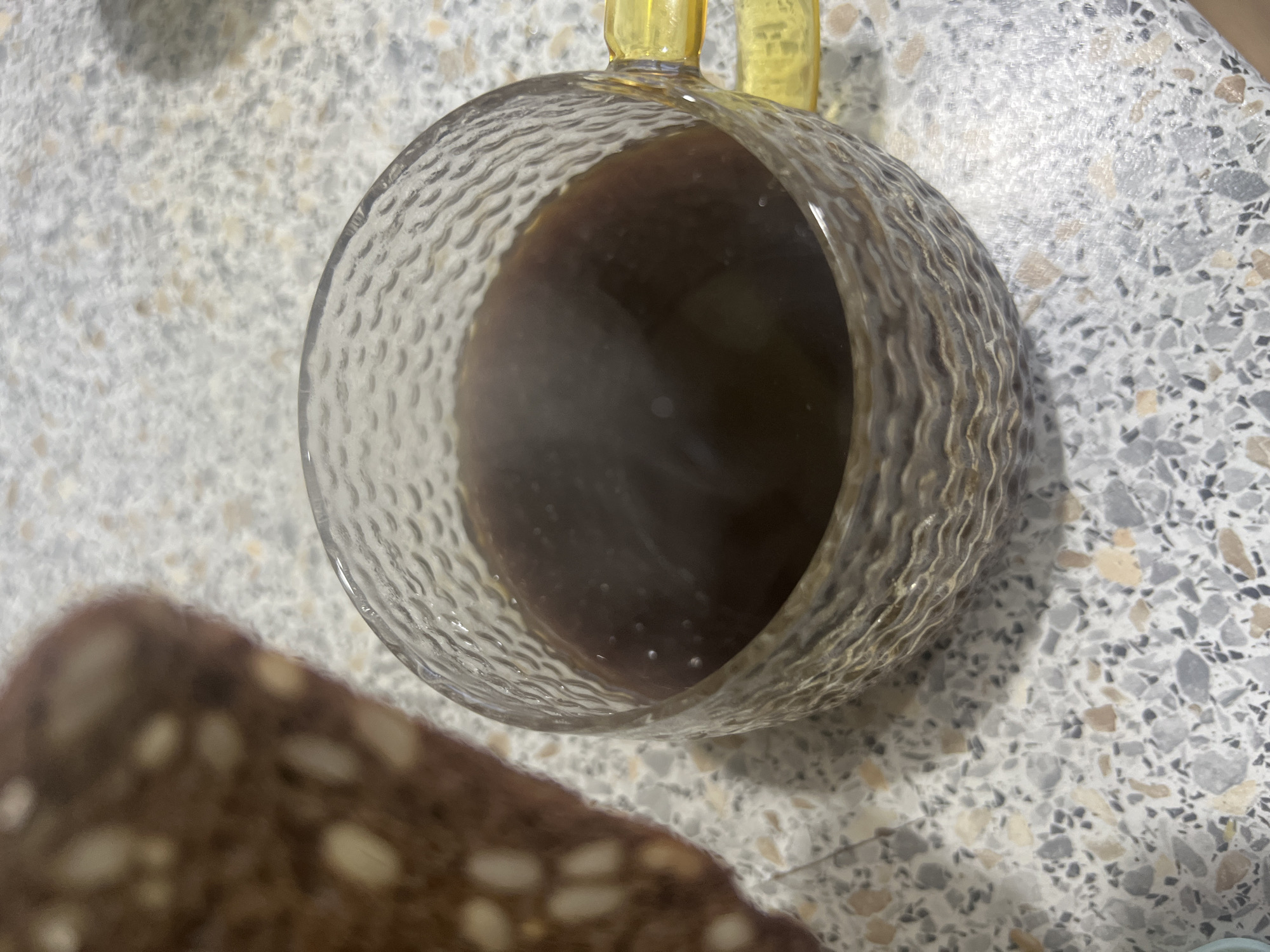 An overhead, close-up shot depicts a clear, textured glass mug holding a dark, steaming beverage, likely coffee or tea. Visible steam rises from the dark liquid, indicating its heat, and small bubbles are present on its surface. The mug has a distinctive rippled or wavy pattern on its exterior and features a bright yellow handle, part of which is visible at the top.
The mug is placed on a light-colored countertop or table with a speckled pattern, resembling terrazzo or granite, composed of various shades including dark grey, light grey, and subtle beige fragments. Partially visible and blurred in the lower-left foreground is a dark piece of bread or baked good, appearing coarse and containing lighter-colored inclusions, possibly seeds or nuts.
The image captures a quiet moment of refreshment or a meal, highlighting the warmth of the drink and the textural elements of the objects. There is no visible text in the image. This indoor scene is located in Chaikovsky, Russia.