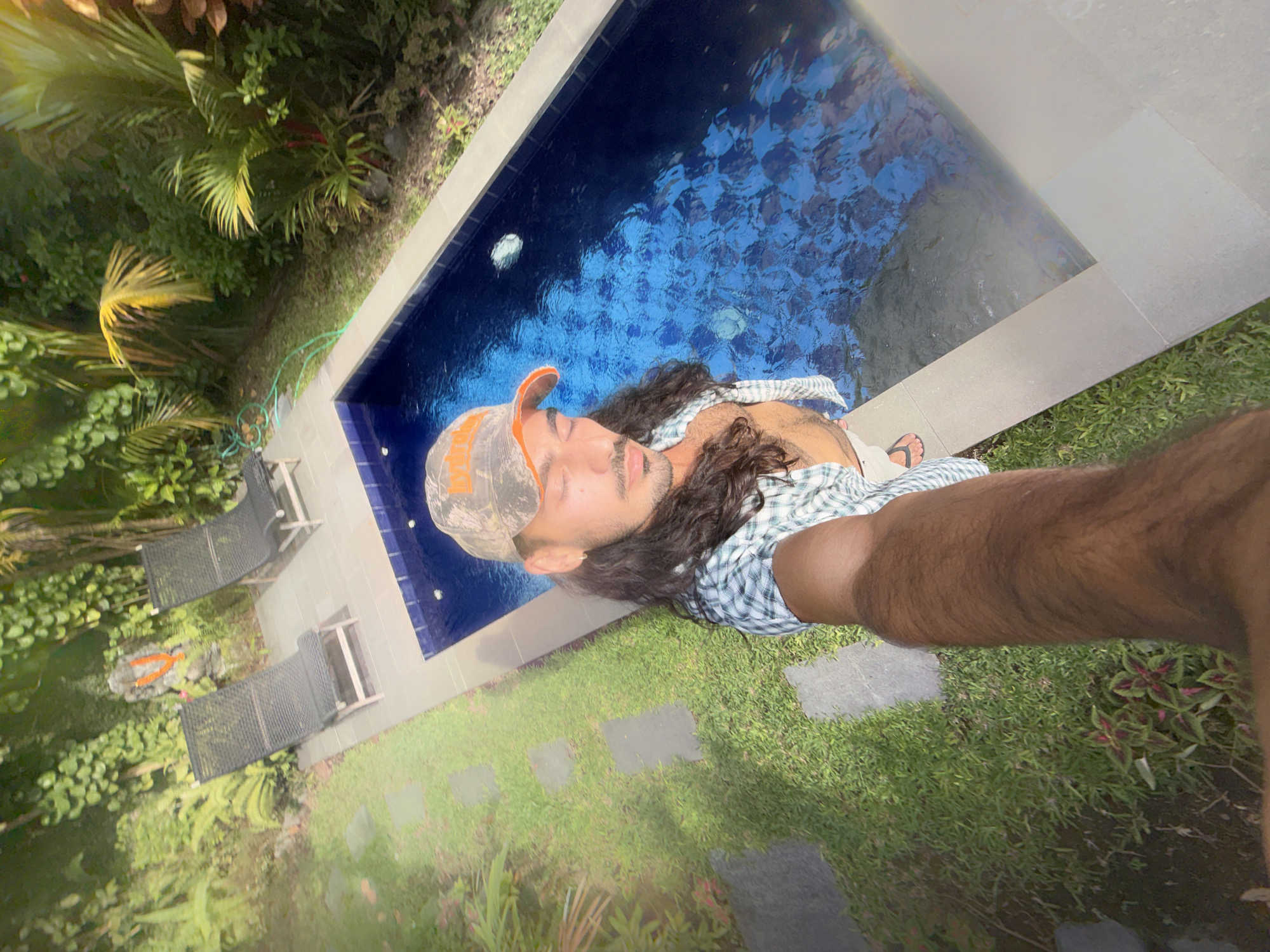A vibrant outdoor selfie captures a person relaxing by a swimming pool in North Kuta, Indonesia, under bright daylight. The image is taken from an overhead, rotated perspective, offering a dynamic view of the scene.
The individual, appearing male, has long dark hair and is wearing a camouflage baseball cap with partially visible orange text, and a green and white plaid shirt worn open. They are positioned by the edge of a rectangular, dark blue-tiled swimming pool, with their body extending into or along the water. Their face is visible, with eyes appearing closed or looking down, conveying a relaxed state. A hairy arm, holding the camera, and a leg with black flip-flops are prominent in the foreground.
The surrounding environment is lush and tropical, featuring vibrant green foliage, palm trees, and various leafy plants. Adjacent to the pool are grey tiled areas and patches of green grass interspersed with dark grey stepping stones. Two black lounge chairs are visible amidst the greenery on the left side of the frame. The water in the pool shows ripples and reflections, indicating clear and sunny weather. The overall scene suggests a leisurely moment in a tropical outdoor setting.