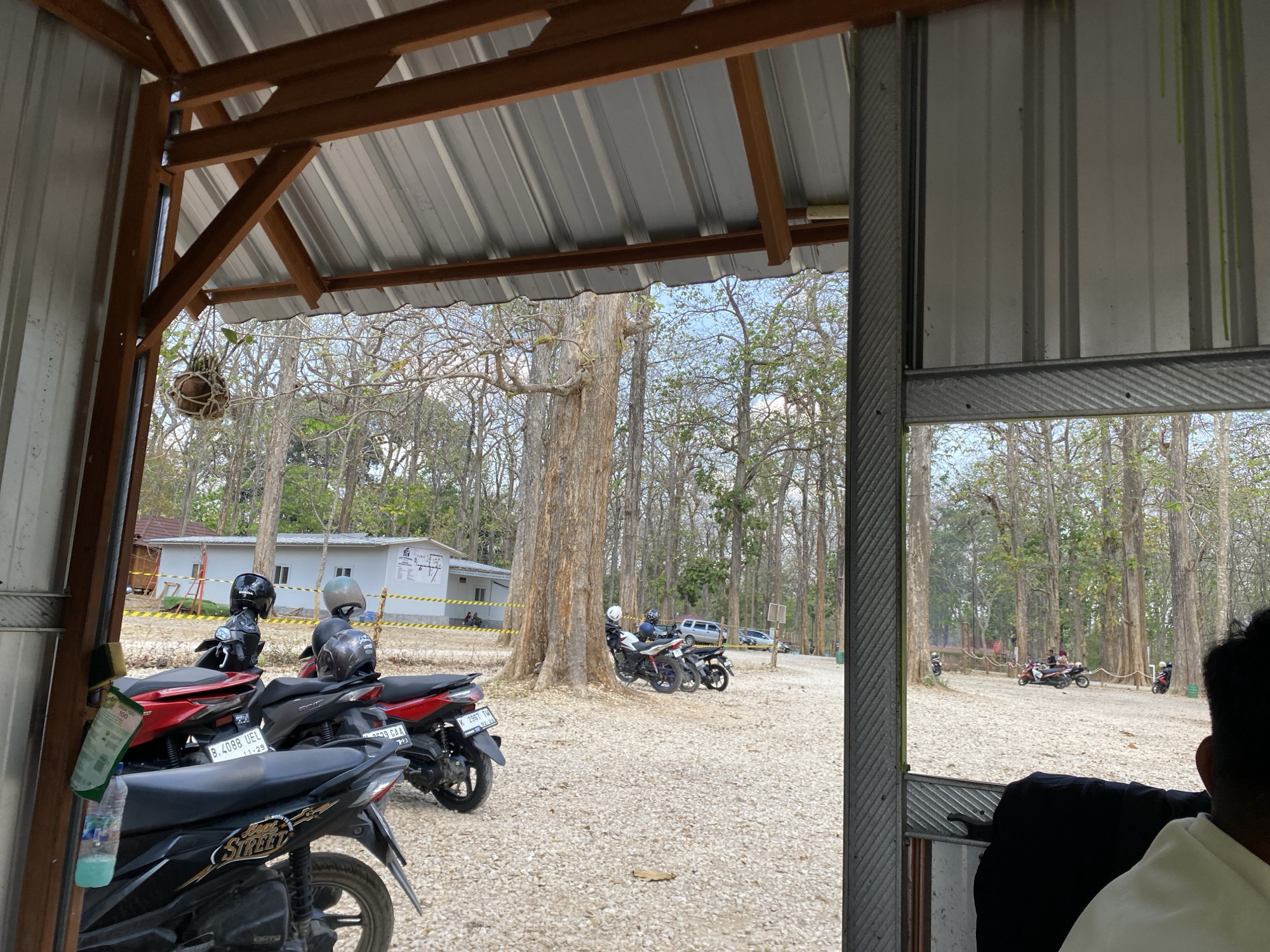 The image depicts an outdoor scene viewed from under a covered structure, likely a carport or open-air shelter. The structure has a corrugated metal roof supported by wooden beams and metal framing. In the foreground, several motorcycles are parked on a gravel surface. One motorcycle in the left foreground has a bottle of liquid and a green leaflet attached to its handlebars. The background shows a dry, wooded area with numerous trees, many of which appear to be deciduous and have lost their leaves. A white building with a yellow stripe is visible in the middle distance, and several people and motorcycles are scattered throughout the scene, suggesting a parking area or recreational spot. The lighting indicates it is daytime, with a clear sky visible between the tree branches. The overall impression is one of a rural or semi-rural location, possibly a nature park or roadside rest area.