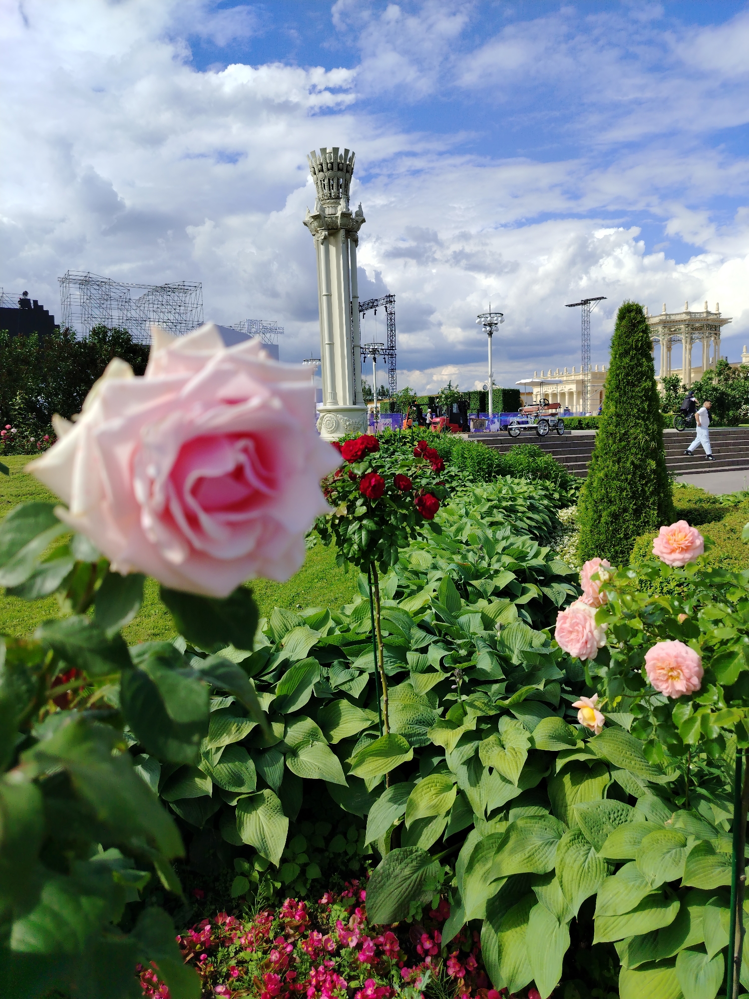 The image depicts a sunny day at VDNKh (Exhibition of Achievements of National Economy) in Moscow, Russia. In the foreground, a soft pink rose is in sharp focus, with a lush green garden bed of hosta plants and smaller pink flowers below. To the right, a tall, conical evergreen tree stands prominently. In the mid-ground, red roses are visible, and a person in white attire is walking up a set of stairs. The background features a classical archway, part of the VDNKh pavilions, and several large metal structures that appear to be stages or scaffolding, possibly for an event. The sky is a vibrant blue with scattered white clouds.