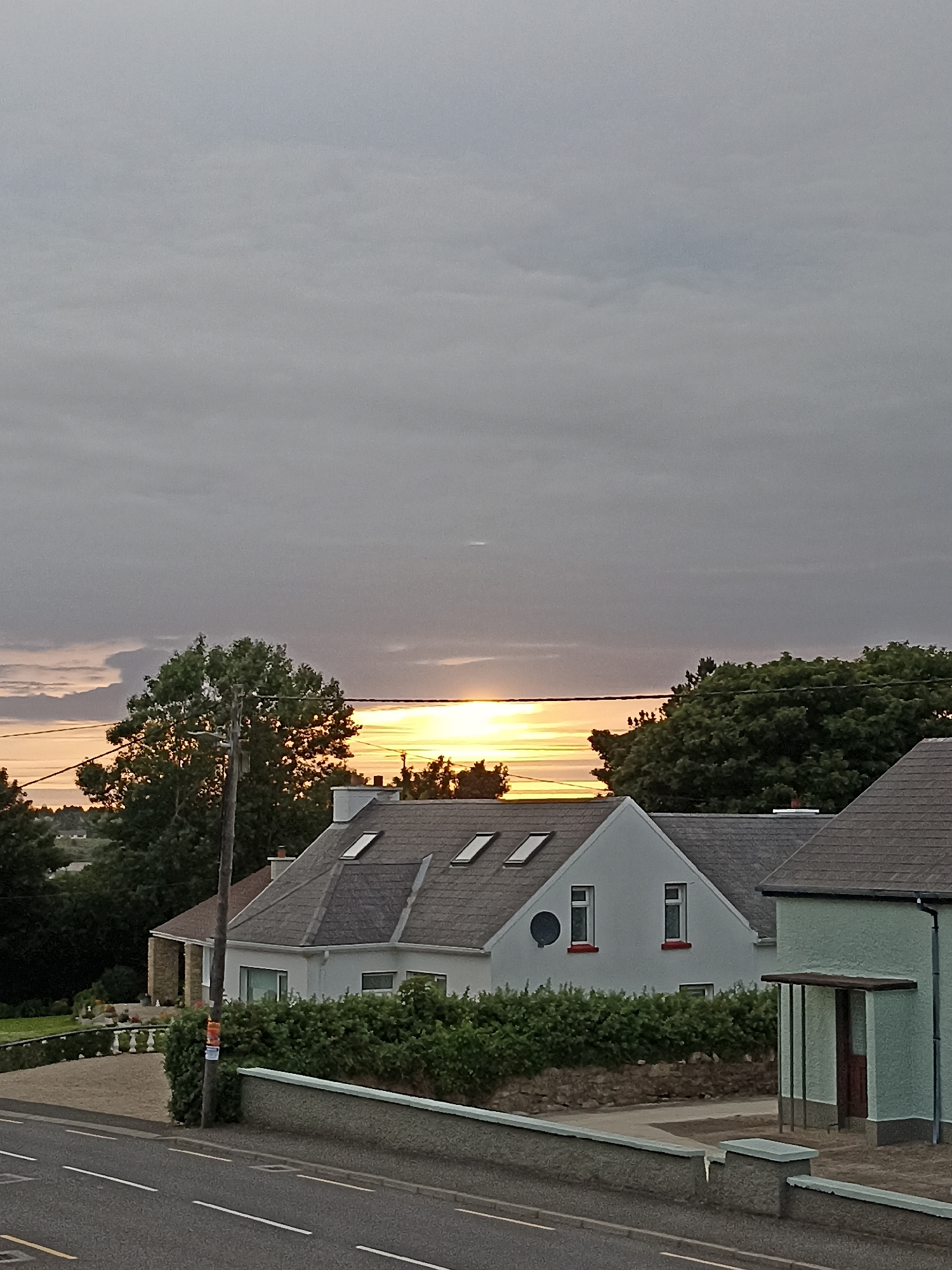 The image shows a view of Annagry, Ireland, at dusk. The sky is overcast with a band of bright, orange light visible on the horizon, suggesting the sun is setting. In the foreground, there are several houses with pitched roofs, some featuring skylights. A road runs along the bottom of the image, with a low stone wall separating it from a manicured garden area and more houses behind. Trees are visible throughout the scene, with a large tree dominating the left side of the frame. There are no people or animals visible.