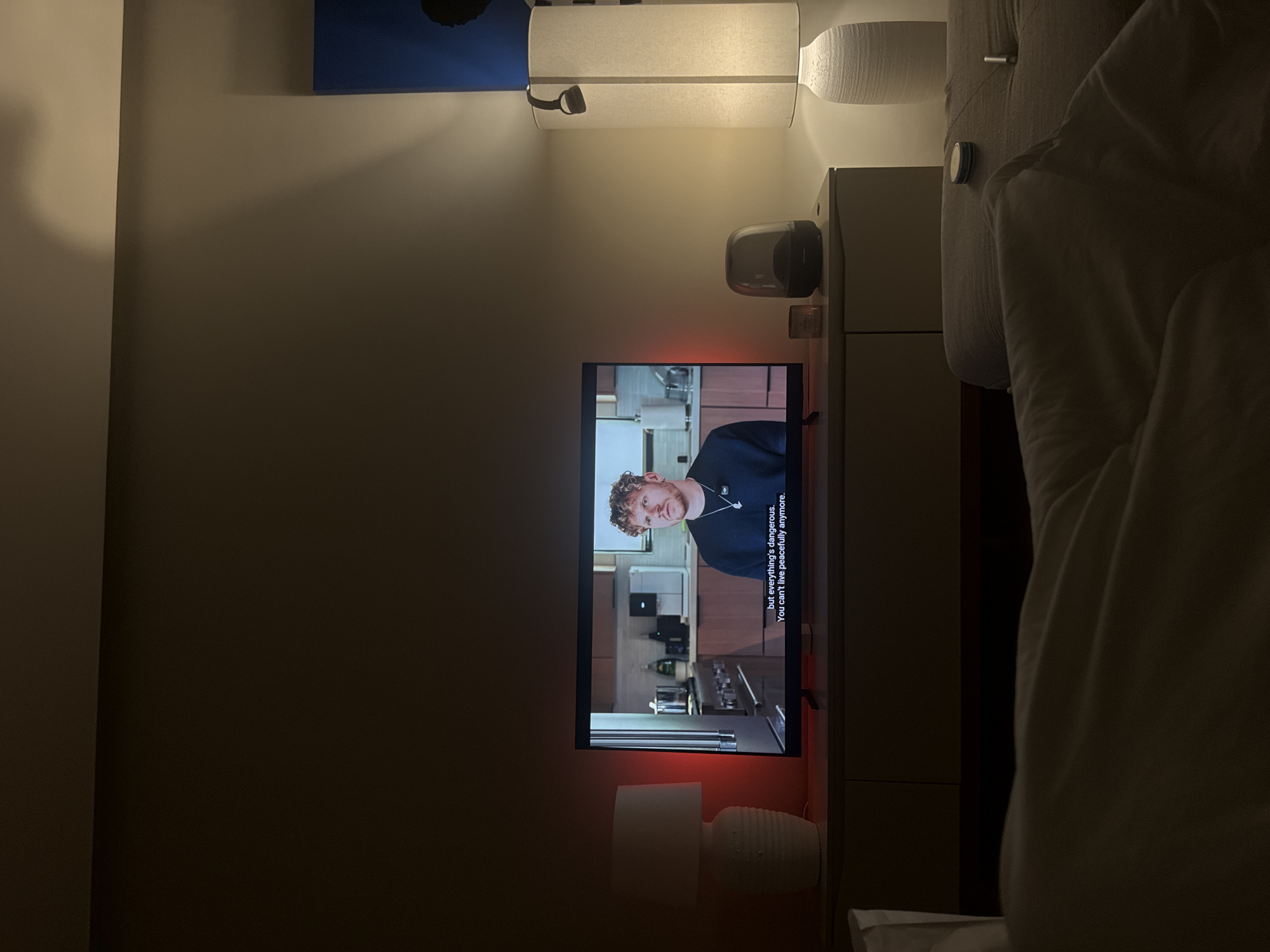 An indoor scene shows a television displaying a man with curly hair, wearing a dark shirt and a necklace. Subtitles read, "but everything's dangerous. You can't live peacefully anymore." The television is placed on a cabinet. To the left of the television is a white table lamp. To the right of the television, on the cabinet, are a small candle and a black and gray speaker. Further to the right is a tall white floor lamp. The foreground shows a bed with white and gray blankets. The overall ambiance is dim, suggesting it is nighttime or the room is intentionally darkened.