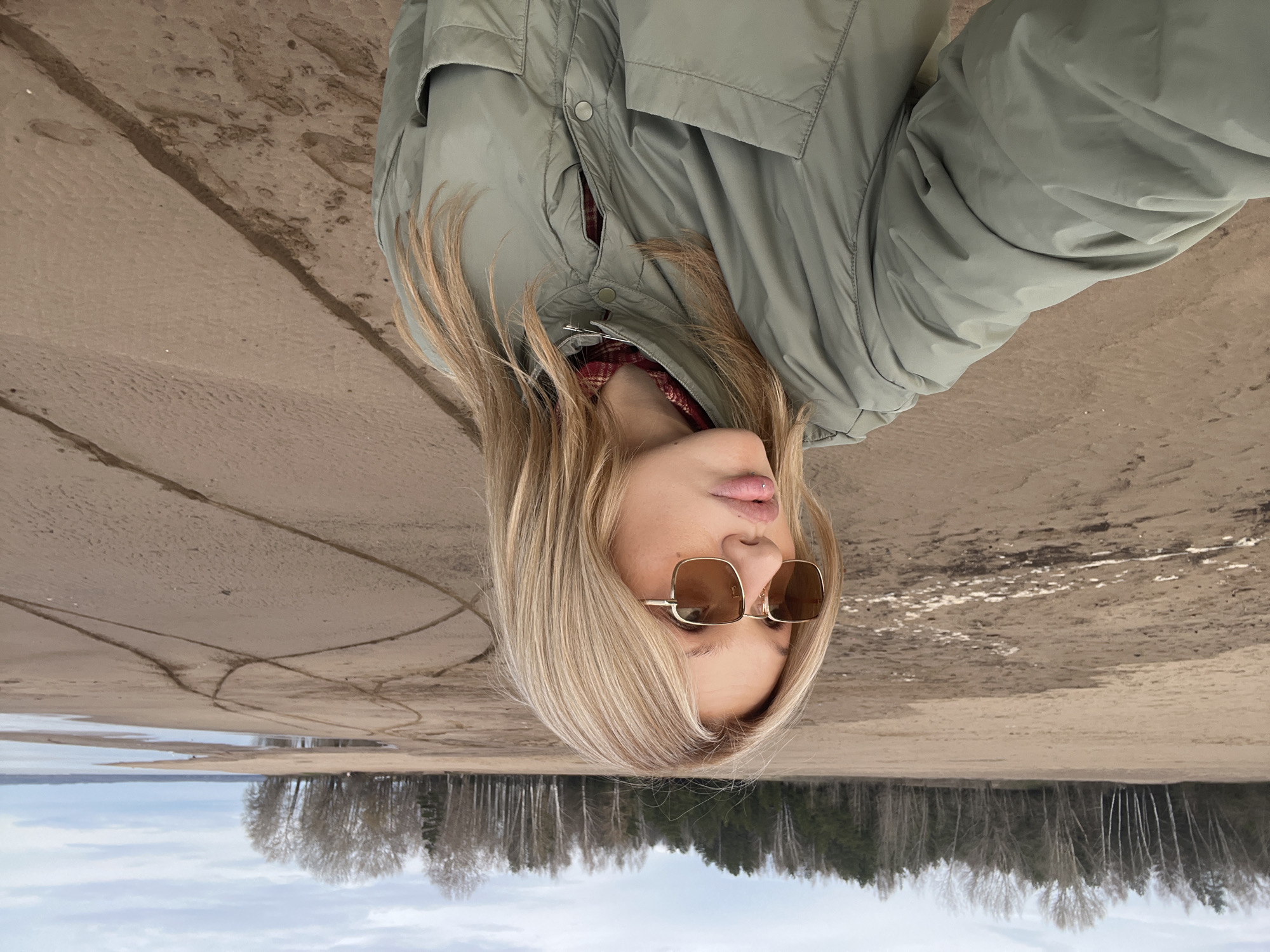 A selfie taken on a beach in Noviy, Russia, features a woman with blonde hair wearing sunglasses and a green jacket, and a red and black flannel shirt. The background consists of a sandy beach transitioning to a forest. The photograph is upside down.