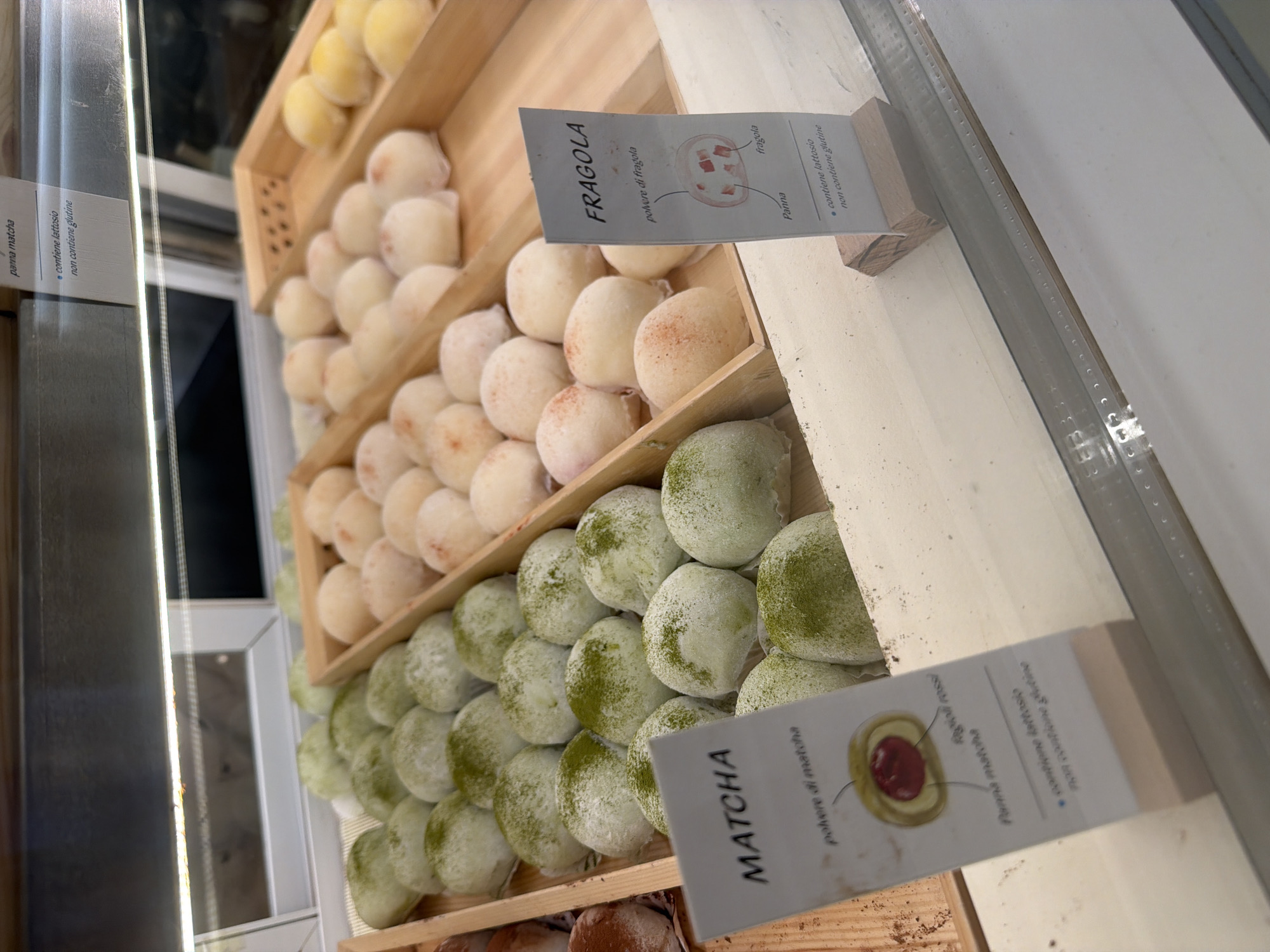 The image shows a display case filled with various mochi. On the left side, there are green mochi, labeled "MATCHA". A sign shows a diagram of the matcha mochi, indicating "Panna matcha" (matcha cream), "polvere di matcha" (matcha powder), and "ragioi rossi" (red filling). The sign also states "contiene lattosio non contiene glutine" (contains lactose, does not contain gluten). To the right, there are light pinkish-white mochi, labeled "FRAGOLA" (strawberry). A sign for the strawberry mochi shows a diagram with "Panna" (cream), "fragola" (strawberry), and "polvere di fragola" (strawberry powder). This sign also indicates that the mochi "contiene lattosio non contiene glutine". In the background, there are more mochi in trays, some appearing to be plain or possibly a different flavor. The display case appears to be made of wood and glass, suggesting it is likely in a bakery or dessert shop. The lighting in the image is bright and artificial, typical of a retail environment. The overall presentation is neat and organized, designed to showcase the confectionery items.
