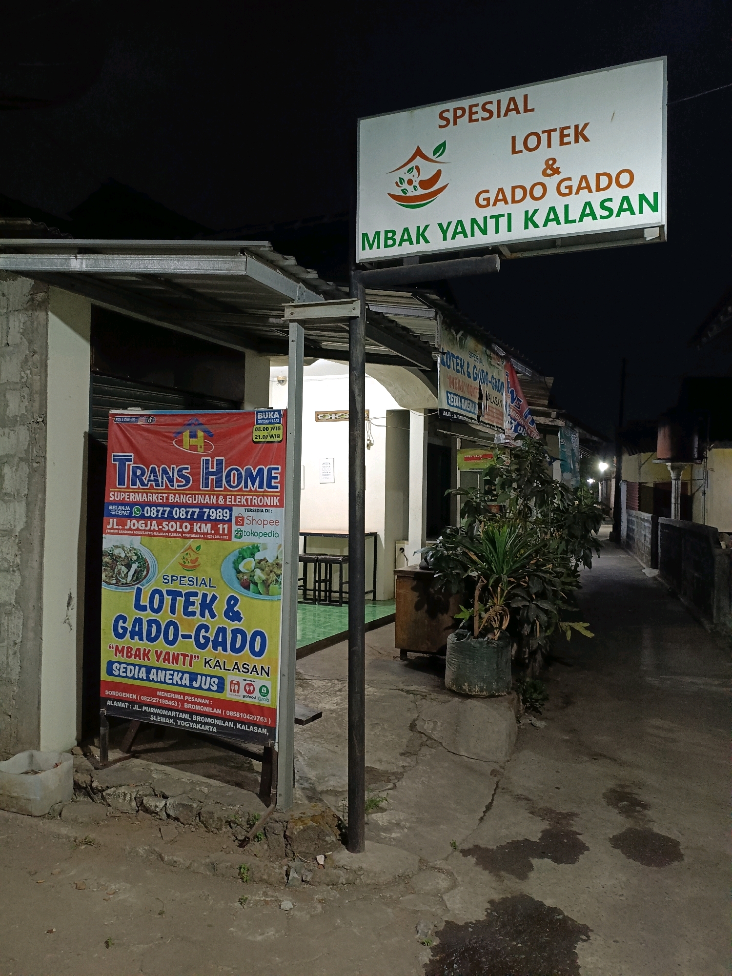 The image depicts a nighttime street scene in Purwomartani, Indonesia, showcasing two prominent signs advertising food services. The primary sign, a large white rectangular board mounted on poles, prominently displays "SPESIAL LOTek & GADO GADO MBAK YANTI KALASAN" in orange and green lettering. It features a logo with a stylized bowl of food and leaves. To the left, a vertical banner-style sign for "TRANS HOME SUPERMARKET BANGUNAN & ELEKTRONIK" is also visible. This banner also advertises "SPESIAL LOTek & GADO-GADO 'MBAK YANTI' KALASAN" with images of the dishes and contact information. The background reveals a row of buildings and a narrow pathway, suggesting a commercial or residential area. The lighting suggests it is after sunset, with artificial lights illuminating the signs.