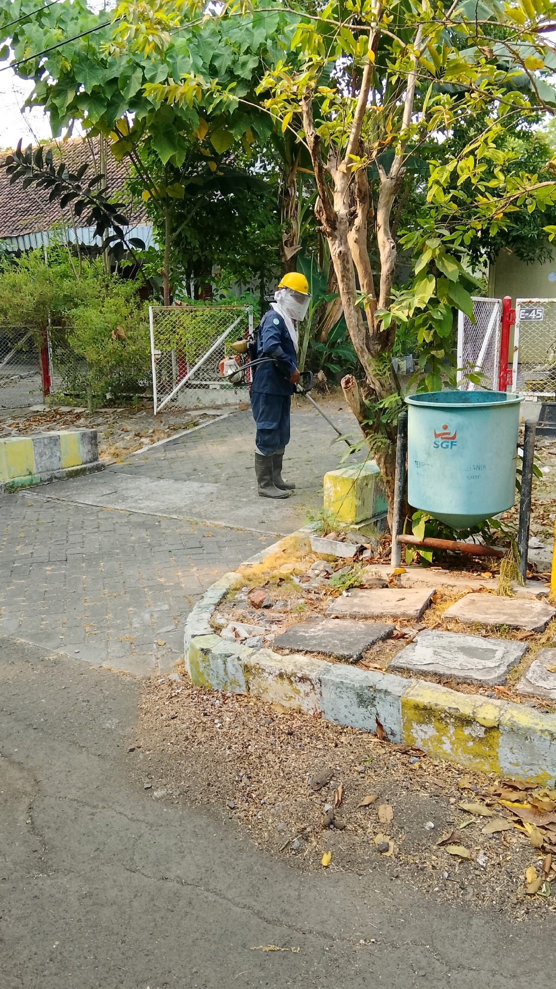 A person, wearing a blue uniform, yellow hard hat, and protective face shield, is operating a string trimmer in an outdoor area. They are positioned in front of a light green trash can with "SGF" logo. The ground is paved with interlocking bricks and edged with concrete barriers painted yellow and grey. The background features lush green trees and foliage, with a glimpse of a building and a white fence. The scene appears to be located in Tlogobendung, Indonesia, given the context.