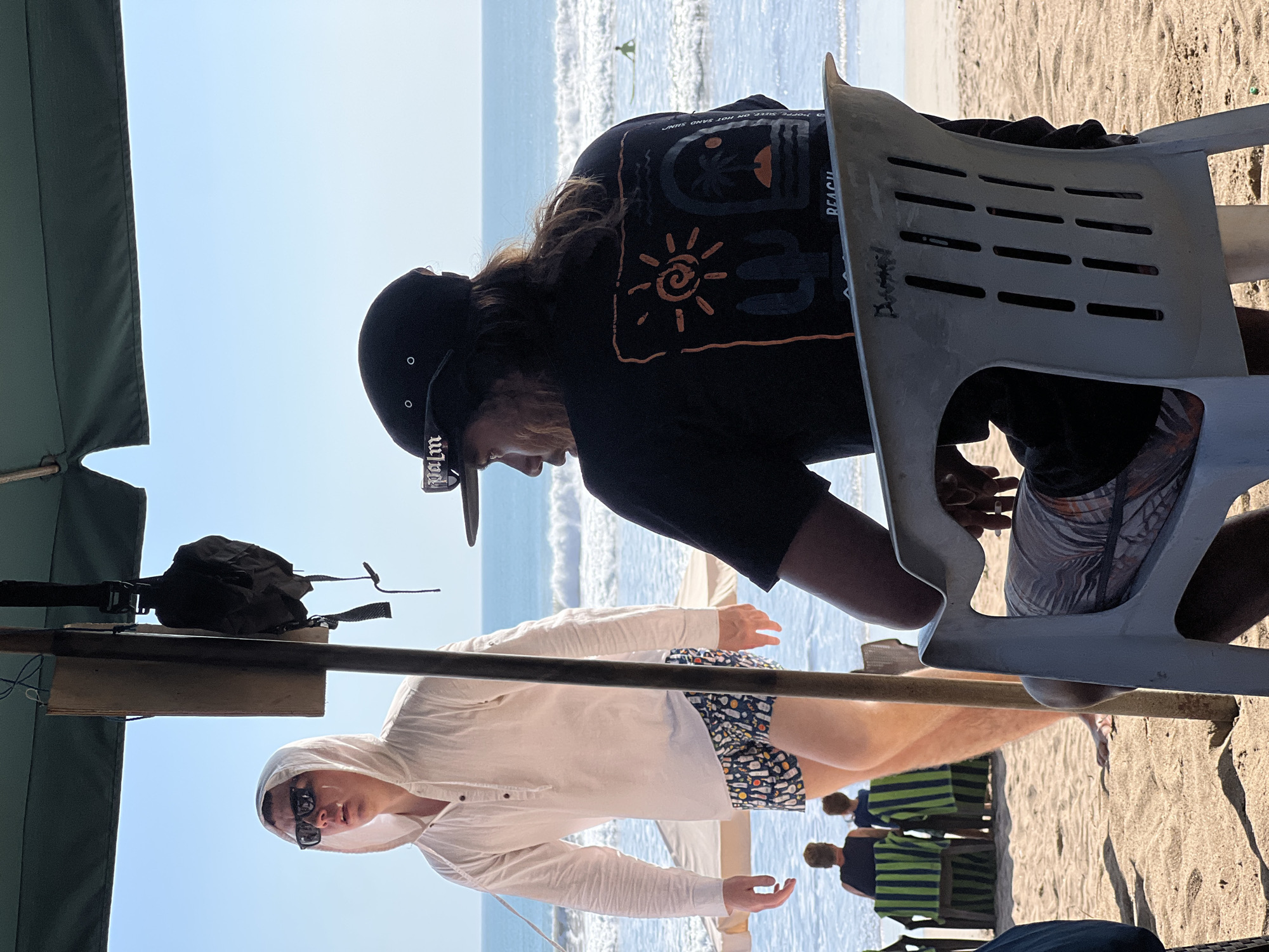In Kuta, Indonesia, on a sunny day at the beach, two men are seated under a green umbrella. The man on the left wears a white hooded shirt and patterned shorts. The man on the right, facing away from the camera, wears a black t-shirt with a graphic design and a black baseball cap. He is seated on a white plastic chair, with a surfing scene depicted on his t-shirt, which also has text that reads "POPPED SURE OR HOT SAND SAND BEACH". The ocean, with small waves, is visible behind them, and two people are seated on lounge chairs in the background. The sand is light brown, and the sky is a clear blue.