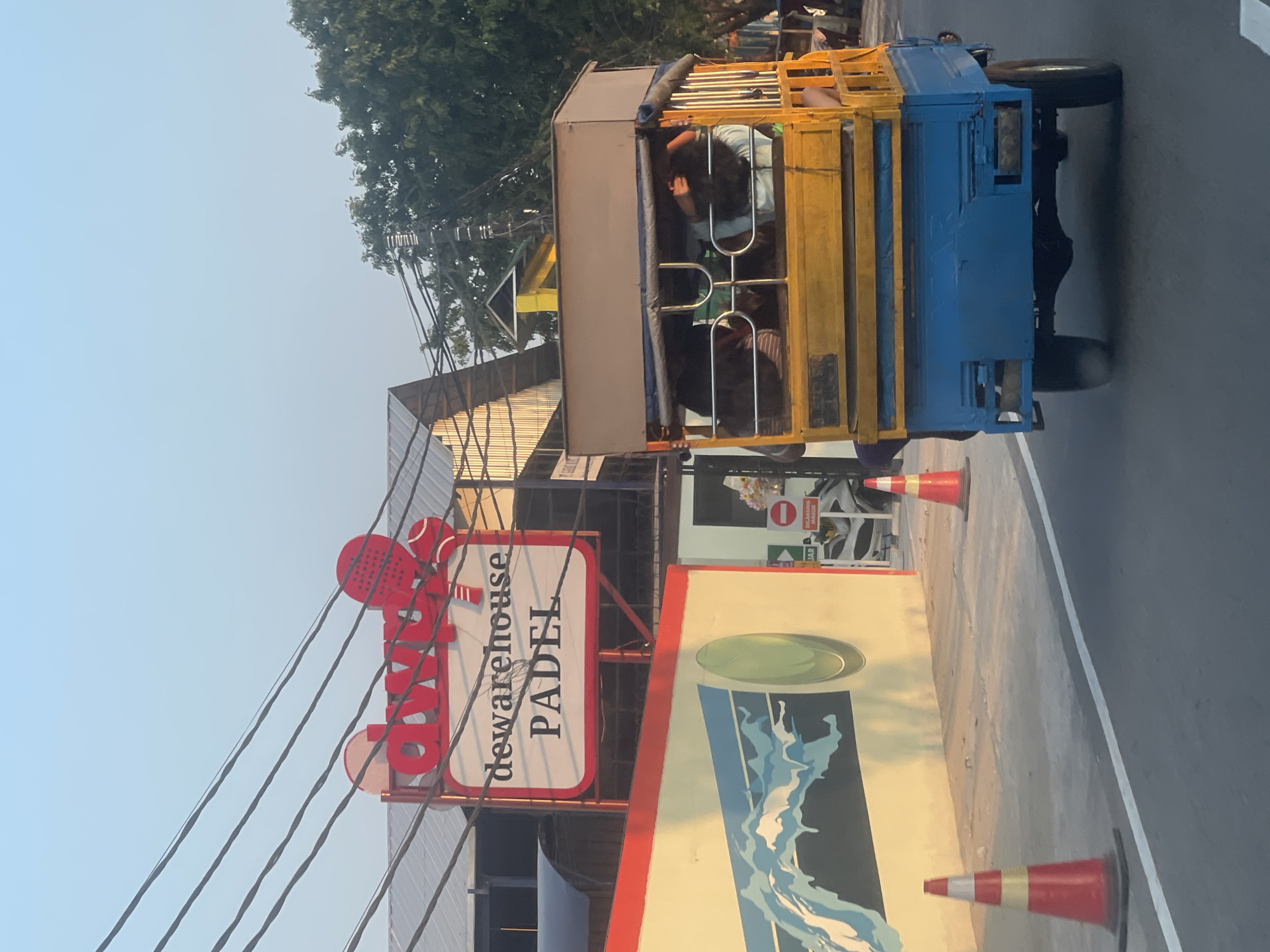 The image shows a bright, sunny street scene, likely in North Jakarta, Indonesia, given the presence of a three-wheeled vehicle often seen in that region. In the foreground, a blue and yellow tuk-tuk, a common mode of transport, is moving along the road. It appears to be carrying several passengers, whose faces are not clearly visible. Two orange traffic cones are placed on the side of the road, suggesting a temporary obstruction or work in progress.
Behind the tuk-tuk, a building with a prominent red and white sign displays the words "dwarehouse PADEL". The sign features stylized graphics of a tennis racket and ball, indicating that the establishment is likely a padel tennis club or facility. The building's architecture includes a metal roof and a mural on an adjacent wall depicting abstract blue waves. Overhead, power lines crisscross the sky, a common sight in urban areas. The overall environment suggests a bustling commercial or recreational area on a clear day.