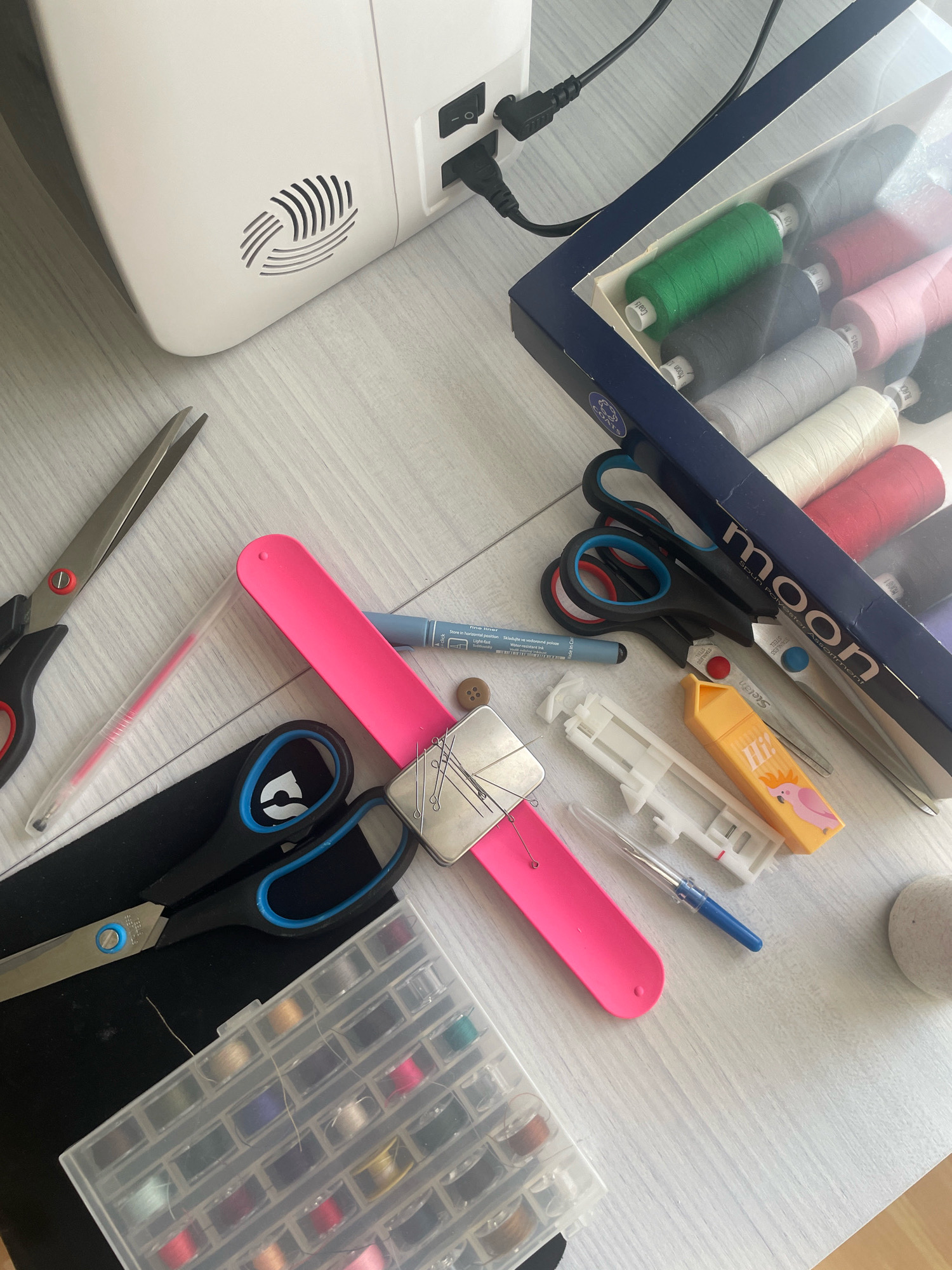 The image depicts a collection of sewing supplies arranged on a light-colored wooden surface, likely a table. In the background, a white sewing machine is visible, connected to power cords. In the foreground, an assortment of items includes various pairs of scissors, spools of thread in a clear box, a pink wrist pincushion with pins, a blue marker, a button, a sewing machine foot, a yellow-orange case with a bird illustration, and a small round object resembling a stone or ball of yarn. The overall impression is that of a well-organized crafting or sewing area. The image provides no information about the location in Atarfe, Spain.