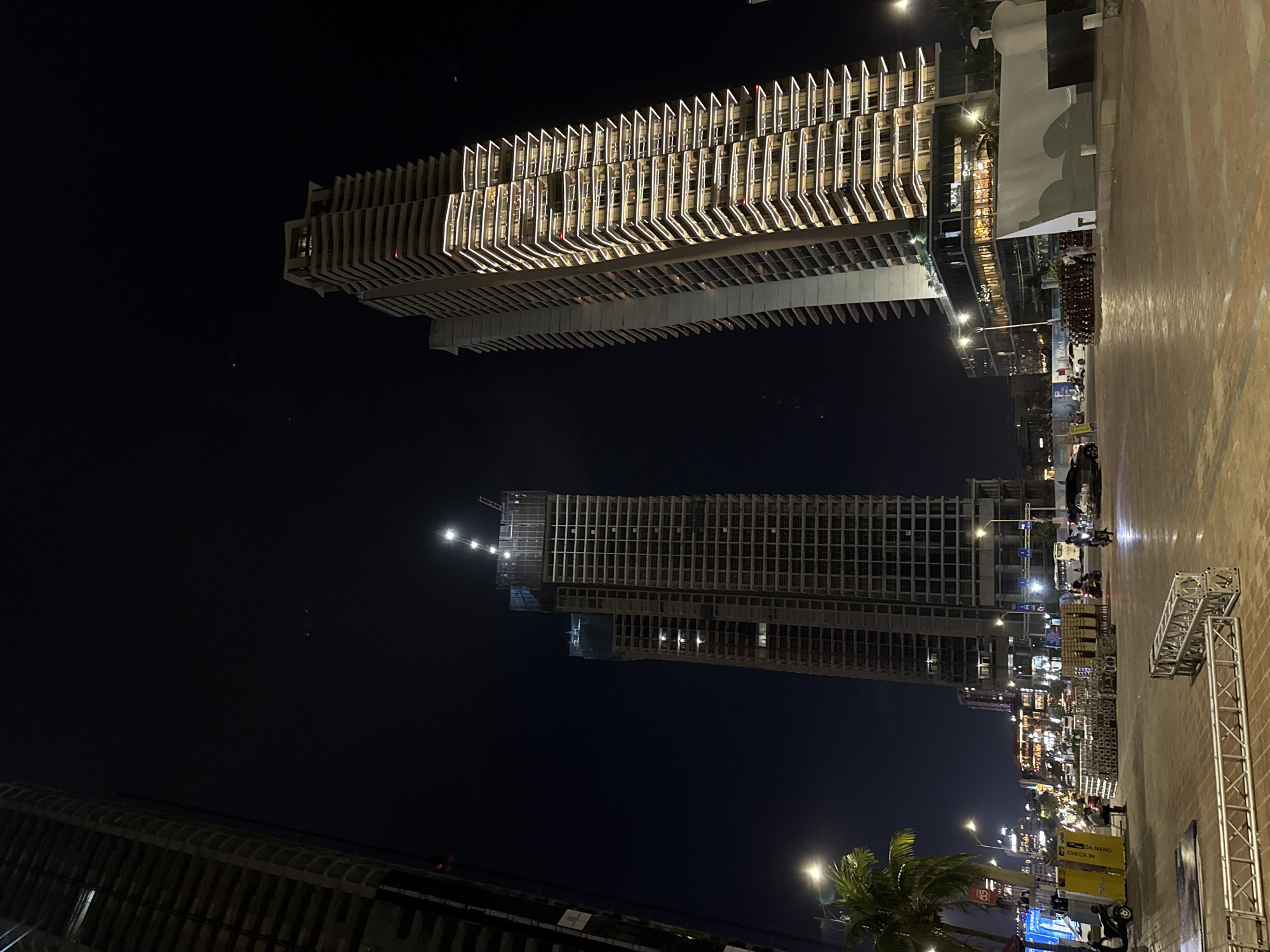 This is a nighttime vertical photograph, depicting an urban scene in what appears to be Đà Nẵng, Vietnam. The image is dominated by two tall buildings against a dark, clear sky, with street-level activity below.
On the left (or top when viewed upright), a modern high-rise building stands prominently. It features a distinctive, illuminated facade composed of numerous horizontal, projecting elements that create a ribbed or louvered effect. Its windows reveal brightly lit interiors.
To the right (or bottom when upright), another tall building is visible, appearing to be under construction. Its concrete framework is exposed, showing a grid of window openings, and construction lights are visible near its upper levels. A small crane structure is partially visible atop this building.
At ground level, a lively street or promenade stretches into the distance. Palm trees with fronds lit by artificial light are visible on the right. Streetlights illuminate the area, and various commercial establishments with lit signs line the street. There are several vehicles, including motorbikes and a white truck, and scattered individuals can be seen on the pavement. Large metal truss structures, possibly for an event stage or signage, are stacked on the right side of the foreground. A large white, tent-like structure or temporary facade covers part of a building at street level on the left.
Notable visible text includes "DA NANG CHECK IN" on a yellow sign and "5G" on a blue sign. The overall scene conveys a busy, active urban environment at night.