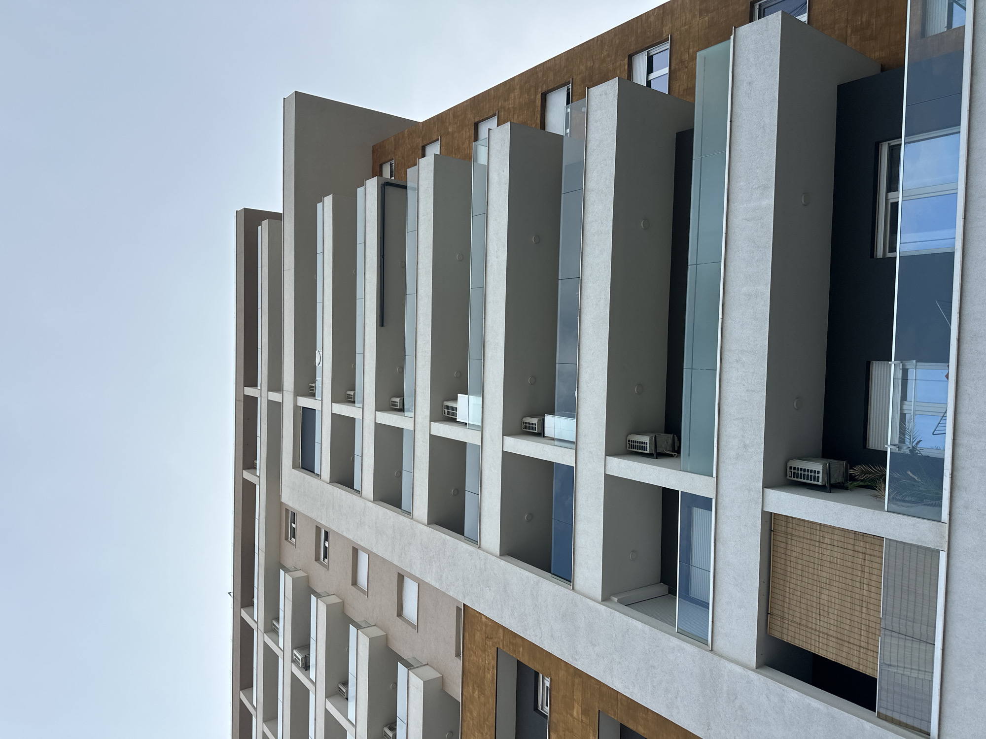 This is a low-angle, Dutch tilt shot of a modern apartment building in Budva, Montenegro, under a clear, overcast sky, suggesting an overcast day. The building features a contemporary architectural design with a facade composed of alternating sections of light grey and a textured brown material. The grey sections are characterized by protruding vertical elements that create recessed balconies or open spaces. These spaces are punctuated by narrow vertical glass panels and often house an air conditioning unit. Several windows are visible, some with blinds, reflecting the muted sky. A bamboo-like blind covers a larger opening in one of the lower balconies. The building appears to be a multi-story residential complex, and the clean lines and geometric shapes suggest a recently constructed or renovated property. There are no people or animals visible in the image. The scene is static and focuses solely on the building's exterior.