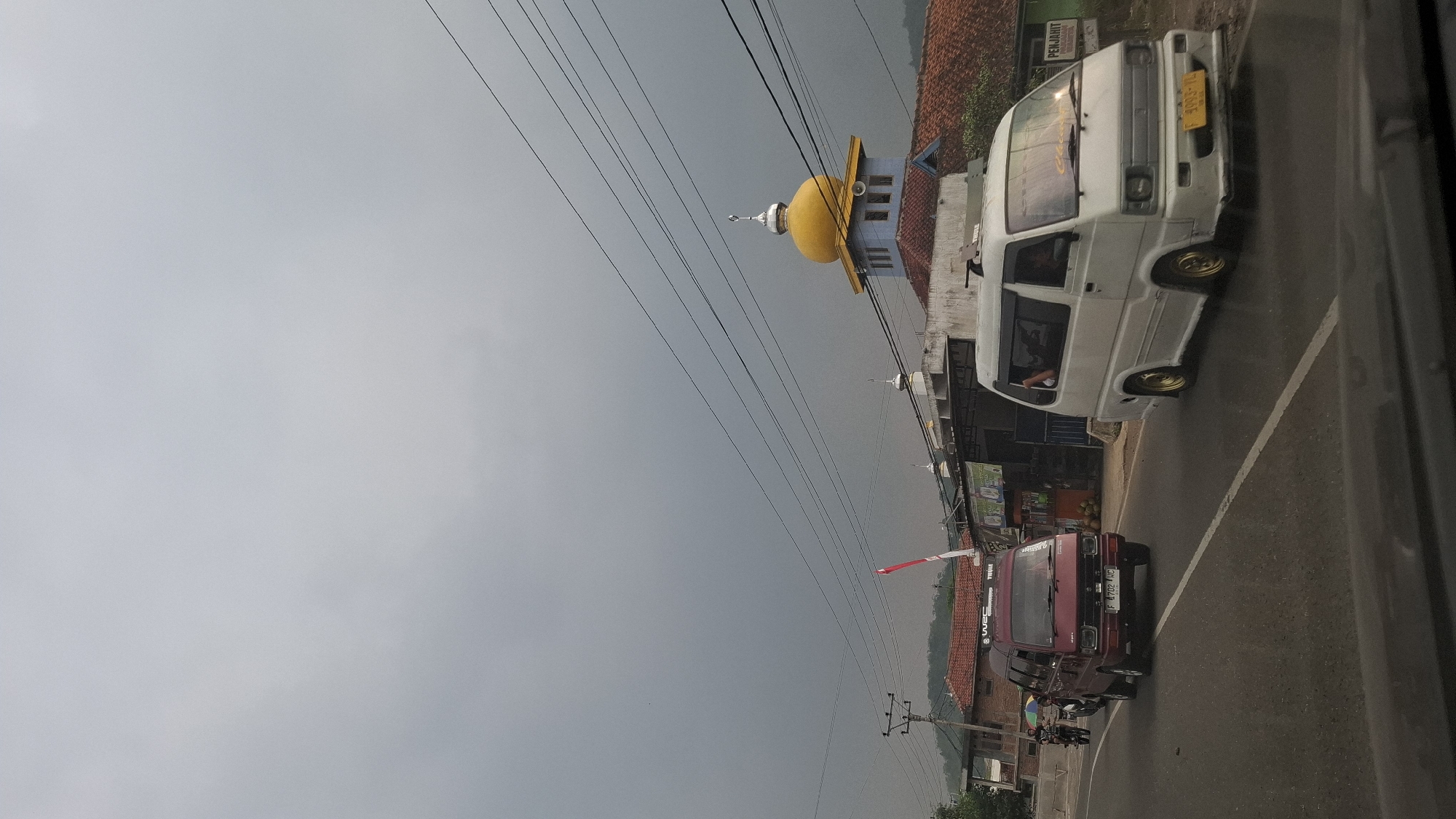 The image captures a roadside scene in Cisalak, Indonesia, under an overcast sky. In the foreground, a dark-colored vehicle, possibly a car, is partially visible at the bottom of the frame, suggesting the photograph was taken from inside another vehicle.
On the road, two vans are prominent. A maroon van, with license plate F 4702 KNC, is in the left lane, moving away from the camera. It has a roof rack. To its right, in the right lane, a white van is also moving away from the camera. This van has a person visible through the driver's side window, and its license plate appears to be F 3993 V. The white van has gold-colored rims.
In the background, a mosque with a distinctive yellow dome is visible. The mosque has blue walls and a red tiled roof. Other buildings with red tiled roofs are also present. A utility pole with electrical wires stretches across the upper portion of the image. A red and white flag is attached to a pole near the mosque.
The overall environment appears to be a developing or residential area, with a mix of commercial buildings and houses along the road. The presence of the mosque and the flag suggests a cultural context within Indonesia. The road appears to be paved with asphalt and has white lane markings. The sky is a uniform gray, indicating either a cloudy day or the approach of rain. A motorcycle with a rider is also visible in the distance, on the left side of the road.