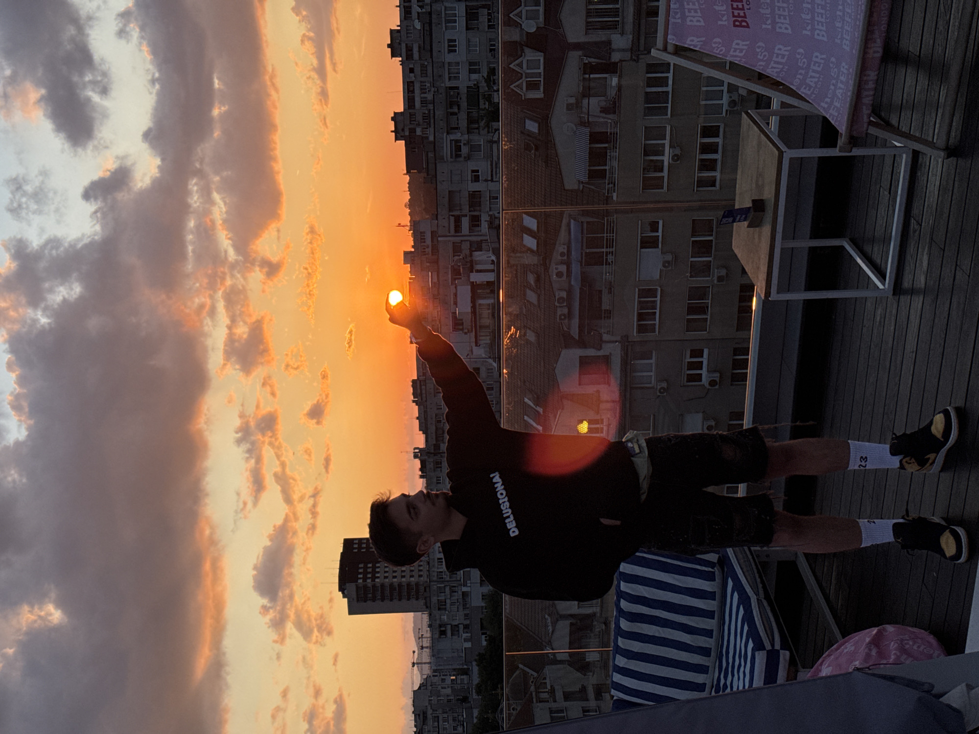 The image depicts a male individual on a rooftop terrace in Sofia, Bulgaria, during sunset. The sky displays vibrant orange and pink hues near the horizon, transitioning to lighter tones and scattered clouds above. The male, facing right and partially silhouetted, is positioned so his right hand appears to hold the setting sun. He wears a black hoodie with "DELUSIONA!" printed in white, dark ripped shorts, white socks featuring the number "23" on the right one, and black and yellow high-top sneakers. A light-colored fanny pack is visible around his waist. The rooftop terrace features dark wooden decking, a clear glass railing, a blue and white striped lounger, a partially visible pink beanbag, a small square wooden table with a white metal frame holding a blue card stand, and a pink and white patterned deck chair with "BEFEATER LONDON" text. The background reveals a dense urban cityscape of Sofia, characterized by numerous multi-story residential buildings and a prominent tall, dark building positioned behind the male's head.