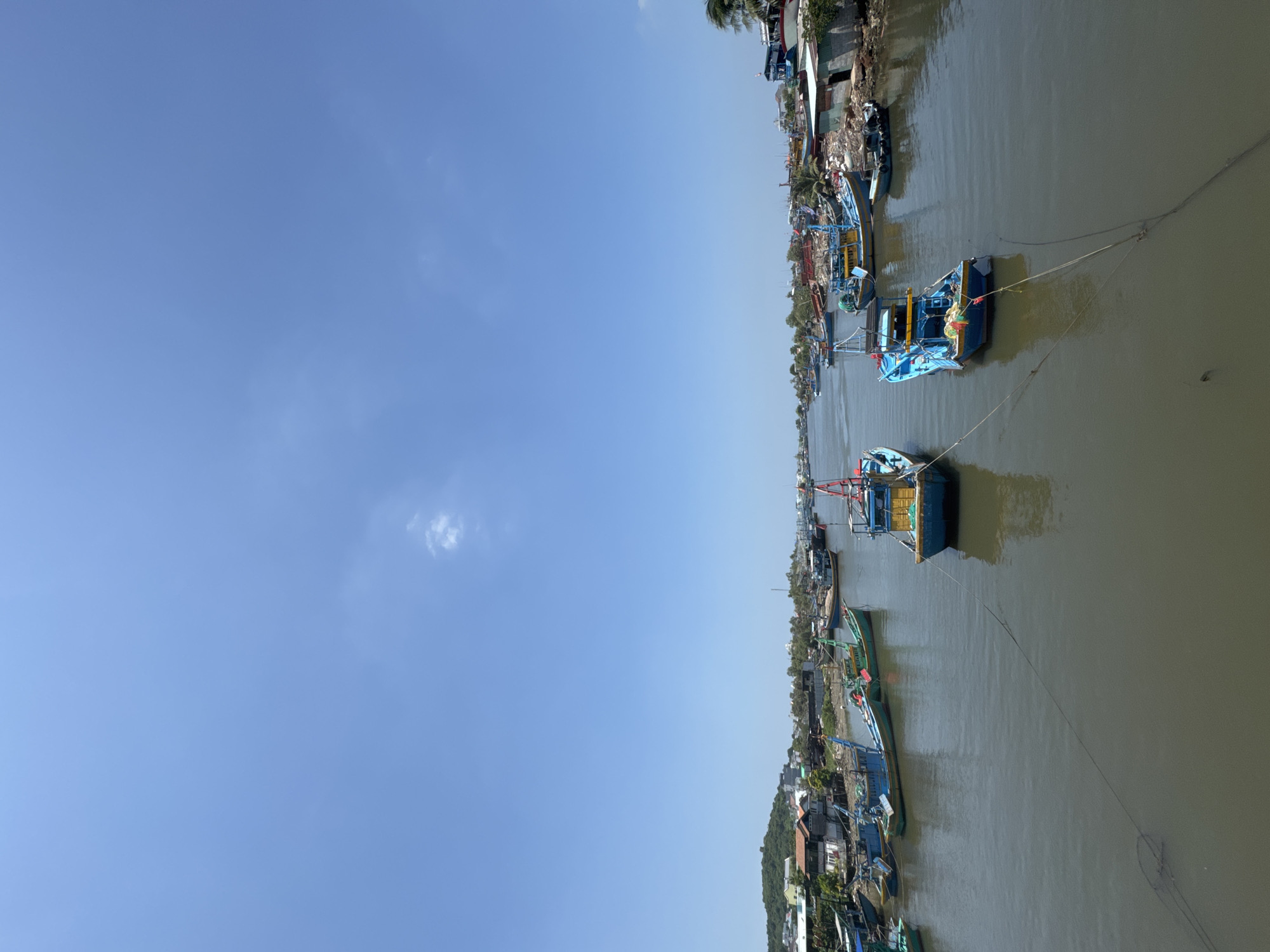 The image depicts a waterway in Phú Thủy, Vietnam, lined with numerous docked fishing boats of varying sizes and colors, predominantly blue and yellow. The boats are moored with ropes to the muddy banks and to each other. On the left side of the waterway, a densely packed area of residential buildings with corrugated metal and tile roofs is visible, nestled against a vegetated hillside. The right bank shows more scattered buildings, some appearing to be small workshops or storage structures, situated on the water's edge with debris and vegetation. The sky is clear and blue with a few wispy clouds. The water is murky and reflects the blue sky. In the foreground, two prominent fishing boats, one blue and yellow and the other predominantly blue, are secured by ropes that extend across the frame. The overall impression is that of a working fishing village or harbor.