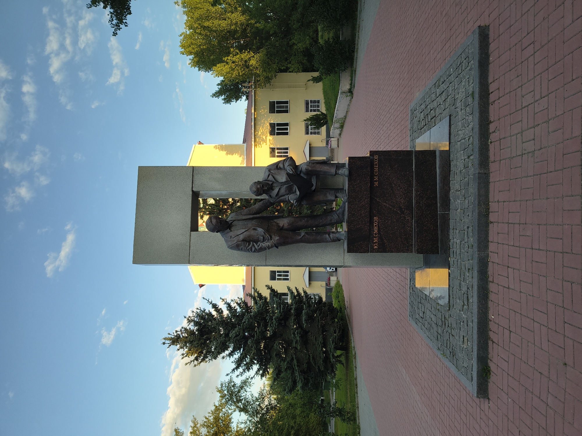 A bronze statue depicting two men stands at the end of a paved walkway in Mikhaylovka, Russia. The man on the left stands upright with his hands in his pockets, while the man on the right is seated, holding a piece of paper. Both men are dressed in suits. The statue is mounted on a dark granite base with inscriptions. Behind the statue is a large, rectangular concrete monument. The walkway is made of red brick pavers. In the background, a yellow building with white-trimmed windows is visible, surrounded by green trees and a blue sky with scattered clouds. A large evergreen tree is positioned to the left of the monument.