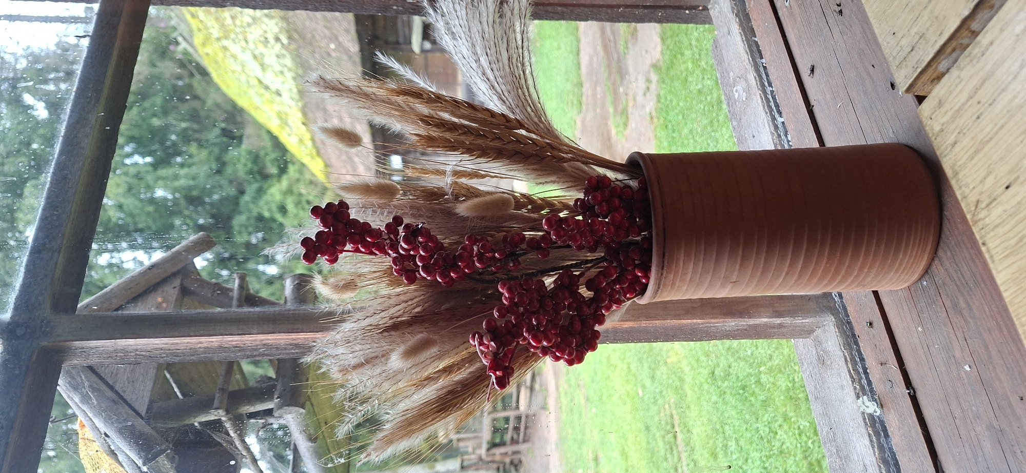 A still life arrangement is positioned on a wooden windowsill. The arrangement consists of a terracotta vase filled with dried wheat stalks, fluffy ornamental grasses, and clusters of bright red berries. The vase is placed on a weathered wooden surface, likely the sill of a window. Outside the window, a green lawn is visible, leading to a rustic building with a thatched roof. Beyond the building, trees are seen, suggesting a rural or natural setting. The windowpane itself shows streaks and water droplets, indicating it may be raining or has recently rained. The overall scene conveys a sense of rustic charm and natural beauty. The location is identified as Saula, Estonia.