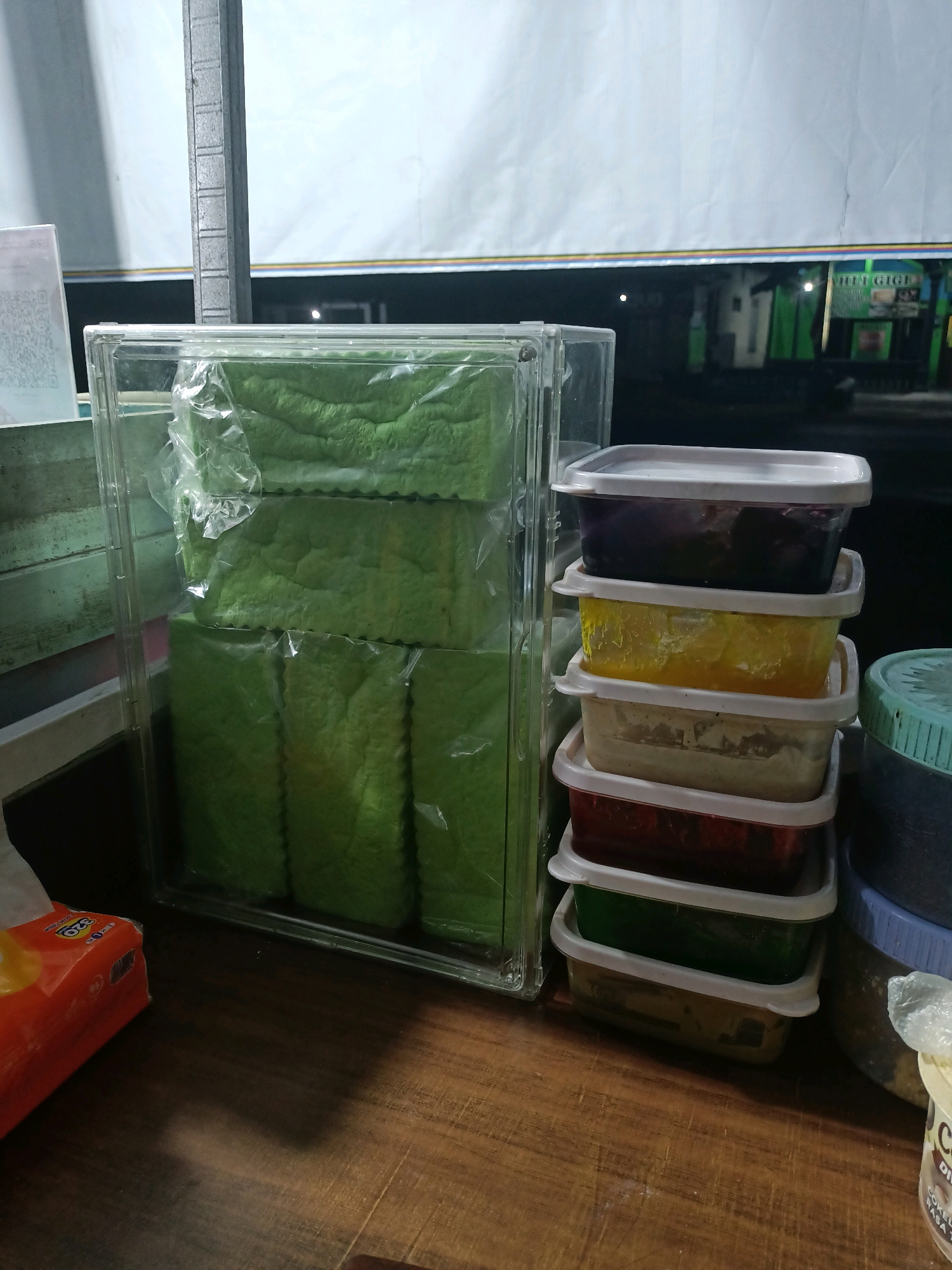 The image shows a close-up of food items displayed on a wooden table, likely in an outdoor stall or shop. In the foreground on the left, a partially opened orange tissue box is visible. To the right of the tissue box is a tall, clear plastic container filled with stacked, rectangular green food items, possibly bread or cake, wrapped in plastic. Beside this, to the right, is a stack of several smaller, white-lidded plastic containers filled with various colored food items, including purple, yellow, beige, red, and green. Further to the right, a large, blue-lidded container is partially visible. In the background, a banner with text, including "HILL GIGI," is stretched across, suggesting a commercial establishment. The overall lighting and setting indicate an evening or nighttime scene. The location is specified as Badas, Indonesia.