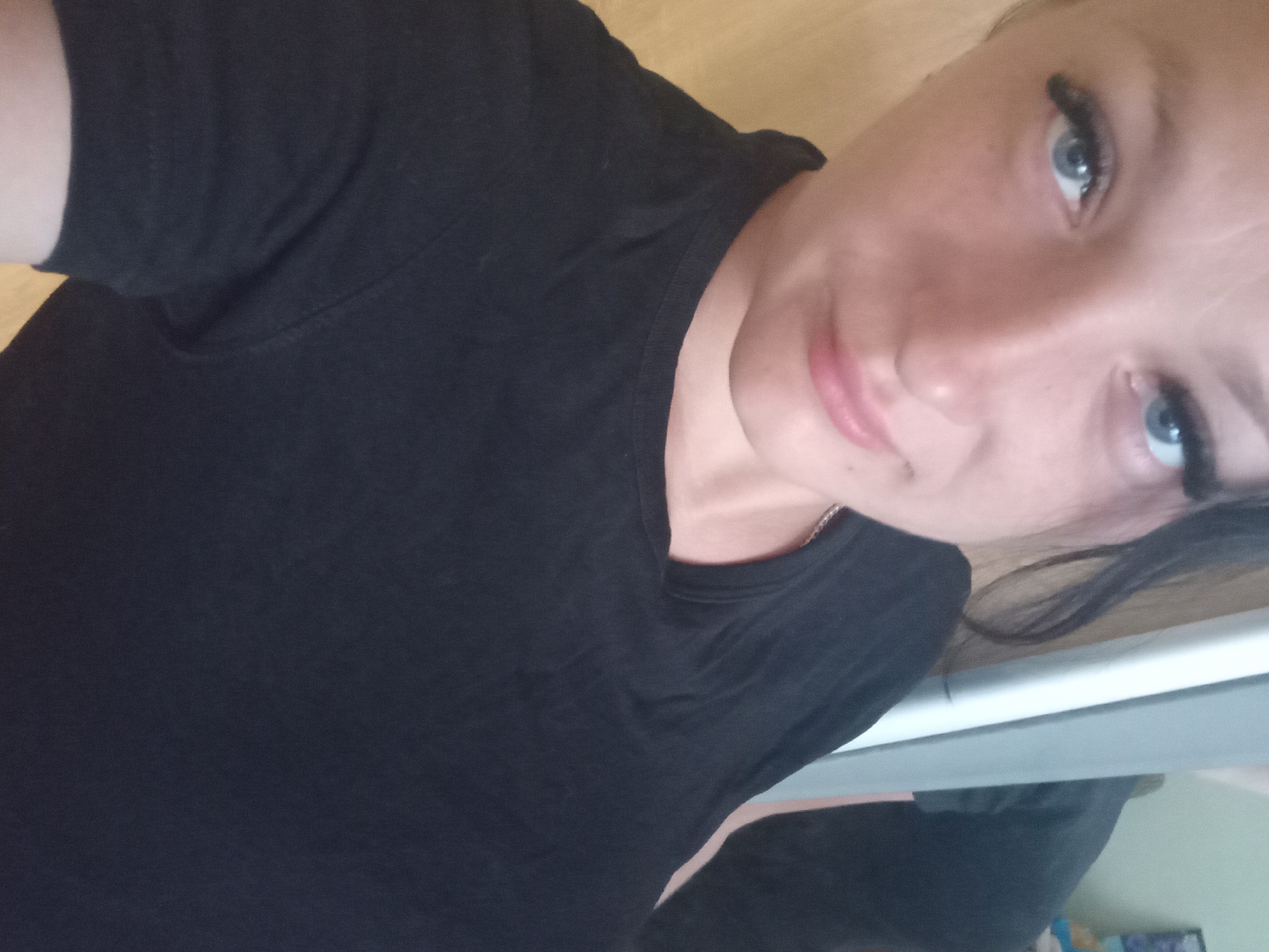 The image is a close-up, low-angle, selfie-style portrait of a woman. She is wearing a black t-shirt and has striking blue eyes and pink lips. Her dark hair is visible, and she appears to be wearing false eyelashes. The background is blurred, showing a light-colored surface that could be a wooden table or floor. The lighting is somewhat dim, casting subtle shadows. The image is oriented sideways, with the woman's face tilted towards the viewer. There is no discernible information in the image that would suggest a location in Chuhuiv, Ukraine.