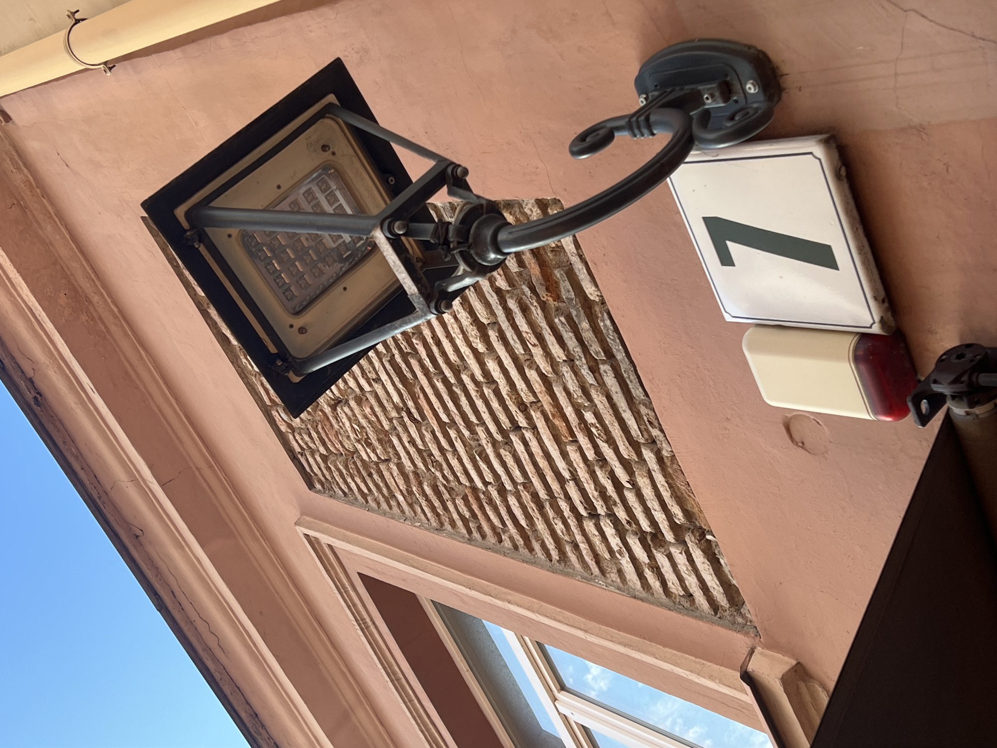 The image shows a building facade in Vilnius, Lithuania. The facade is a salmon pink color and features a large window with a white frame. To the right of the window, a section of the wall is exposed brickwork. A modern black street lamp with an LED light fixture is mounted on the wall. Below the lamp, a white rectangular sign with a black border displays the number "7" in black. To the left of the number sign, a cream-colored rectangular object with a red bottom section is also mounted on the wall. A yellow pipe runs vertically along the right side of the building. The sky is a clear blue.