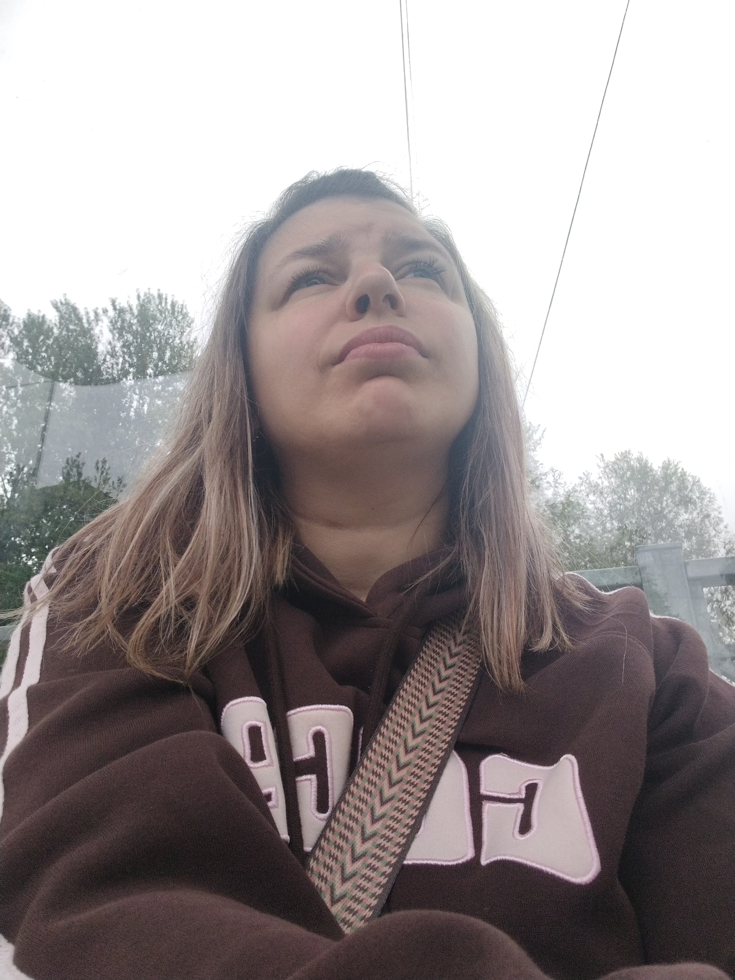 A young woman with long, light brown hair is captured from a low angle, looking upwards. She is wearing a brown hoodie with white stripes on the sleeves and white lettering on the chest. A patterned strap is visible across her chest. The background consists of trees and a bright, overcast sky, suggesting an outdoor setting. The image appears to have been taken on a cloudy day, possibly in a park or natural area within Yaroslavl, Russia, given the context provided. The woman's expression is thoughtful or contemplative.