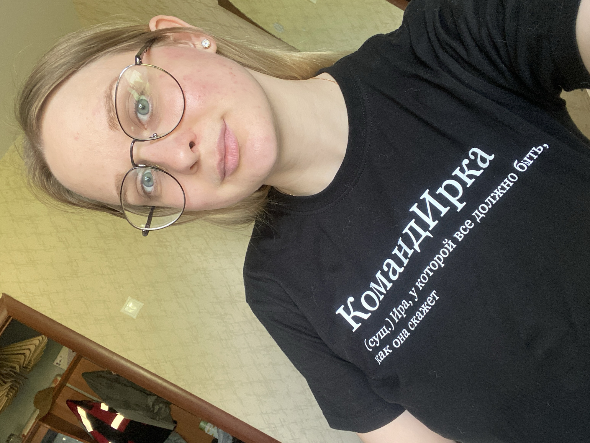 A young woman with blonde hair and glasses is posing for a selfie in what appears to be a residential interior. She is wearing a black t-shirt with white text that reads "КомандИрка (сущ.) Ира, у которой все должно быть, как она скажет". The background includes a textured yellow wall and a wooden door frame with a mirror reflecting some clothing items. The lighting is natural, suggesting it is daytime. The woman's expression is neutral, and she is looking directly at the camera.