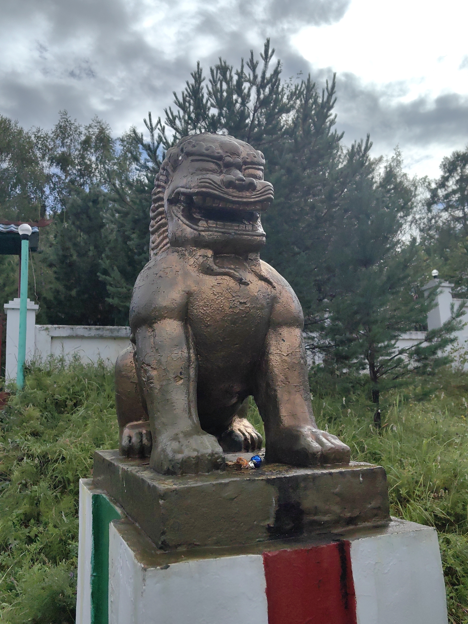 An aged, bronze-colored statue of a Foo dog, also known as a lion-dog, is positioned outdoors. The statue is seated on a multi-tiered pedestal. The Foo dog has a fierce expression with an open mouth displaying teeth and a segmented mane. The bronze finish shows signs of wear and chipping. The pedestal is painted with white, red, and green bands. The statue and pedestal are surrounded by overgrown green grass and foliage. In the background, there are several coniferous trees and a cloudy sky. A white fence and a green pole with a light fixture are visible to the left of the statue. The scene appears to be in an outdoor, possibly park-like setting, and is located in Tibelti, Russia.