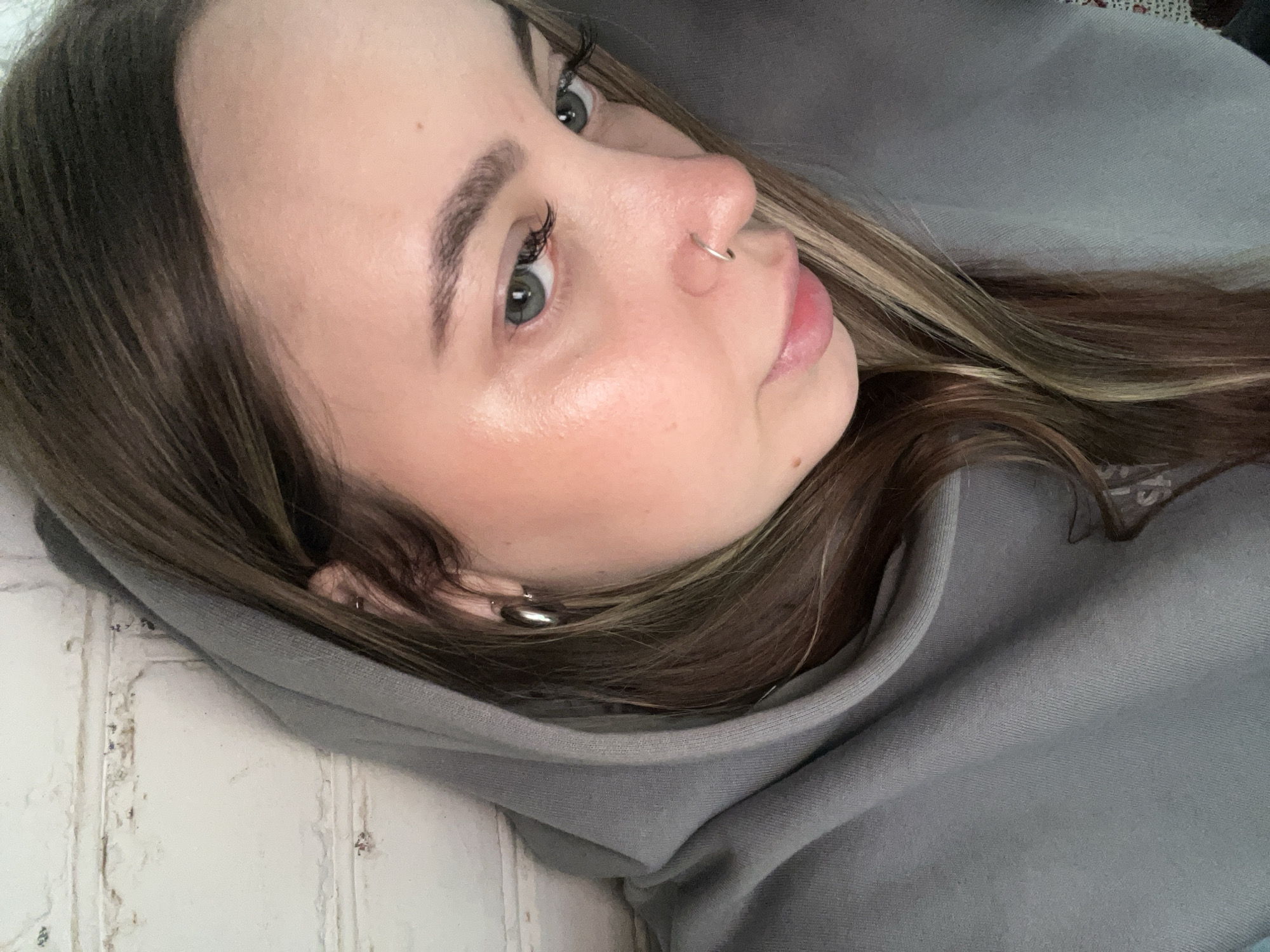 A close-up portrait features a young woman, likely in her late teens or twenties, captured at an oblique angle. She is looking upwards and slightly to her right with a calm and neutral expression. She has fair skin, dark brown hair with lighter brown or blonde highlights, and light-colored eyes, possibly greenish-blue, accentuated by long eyelashes and well-defined eyebrows. A small silver hoop nose ring is visible on her right nostril, and a silver hoop earring adorns her left ear. Her lips have a natural pink tint.
She is wearing a grey hooded sweatshirt, with the hood drawn up around her head, framing her face and providing a soft, textured foreground. On the left side of the image, a white wall with a distressed or brick-like texture is visible, suggesting an indoor setting. The background on the right is a plain grey, likely part of the hoodie or a simple wall. The lighting is soft and even, highlighting the natural contours of her face and giving her skin a slightly dewy appearance. The image shows no visible text. This indoor scene is situated in Novopokrovskaya, Russia.