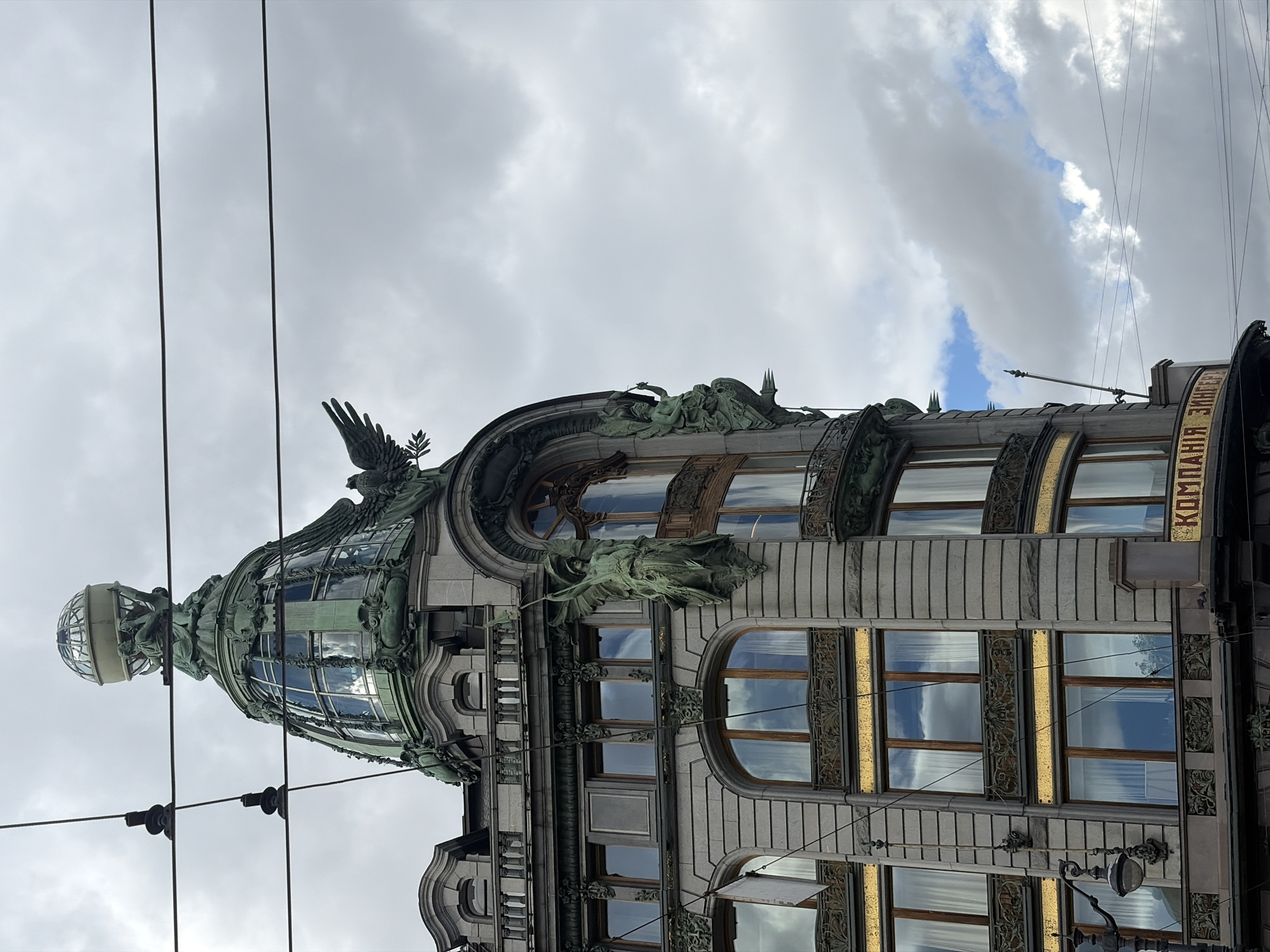 The image captures a grand, ornate building in an urban setting, likely in Saint Petersburg, Russia, given the provided context. The building, viewed from a street level looking upwards, showcases a distinctive Art Nouveau architectural style.
The primary subject is the multi-story facade of the building, characterized by intricate detailing and a prominent domed structure at its pinnacle. The facade is adorned with elaborate dark metal (likely bronze or cast iron) sculptures and decorative elements. Visible sculptures include winged figures, some resembling eagles or angels, positioned at various levels and around the main dome. The dome itself is made of glass and metal, topped with a smaller globe held by a figure, adding to its iconic appearance.
The building features numerous windows, many of which are tall and arched, with some showcasing golden-colored decorative panels between floors and around the window frames. The windows reflect the cloudy sky, suggesting the weather conditions. The main material of the building appears to be light-colored stone or stucco, contrasting with the dark metalwork.
The sky above is mostly overcast with large, textured clouds, though patches of brighter white and some blue sky are visible, indicating it is daytime. Several dark overhead wires crisscross the upper portion of the frame, suggesting the presence of trolleybuses or trams in the area. A classic street lamp is partially visible in the lower right corner, further grounding the scene in a city environment.
Visible text on a curved sign on the building reads "КОМПАНИЯ ЗИНГЕР" (KOMPANIYA ZINGER), which translates to "Singer Company." This indicates the building is likely the famous Singer House (Dom Knigi) in Saint Petersburg. No human activity is visible, making the building itself the central focus of the photograph.