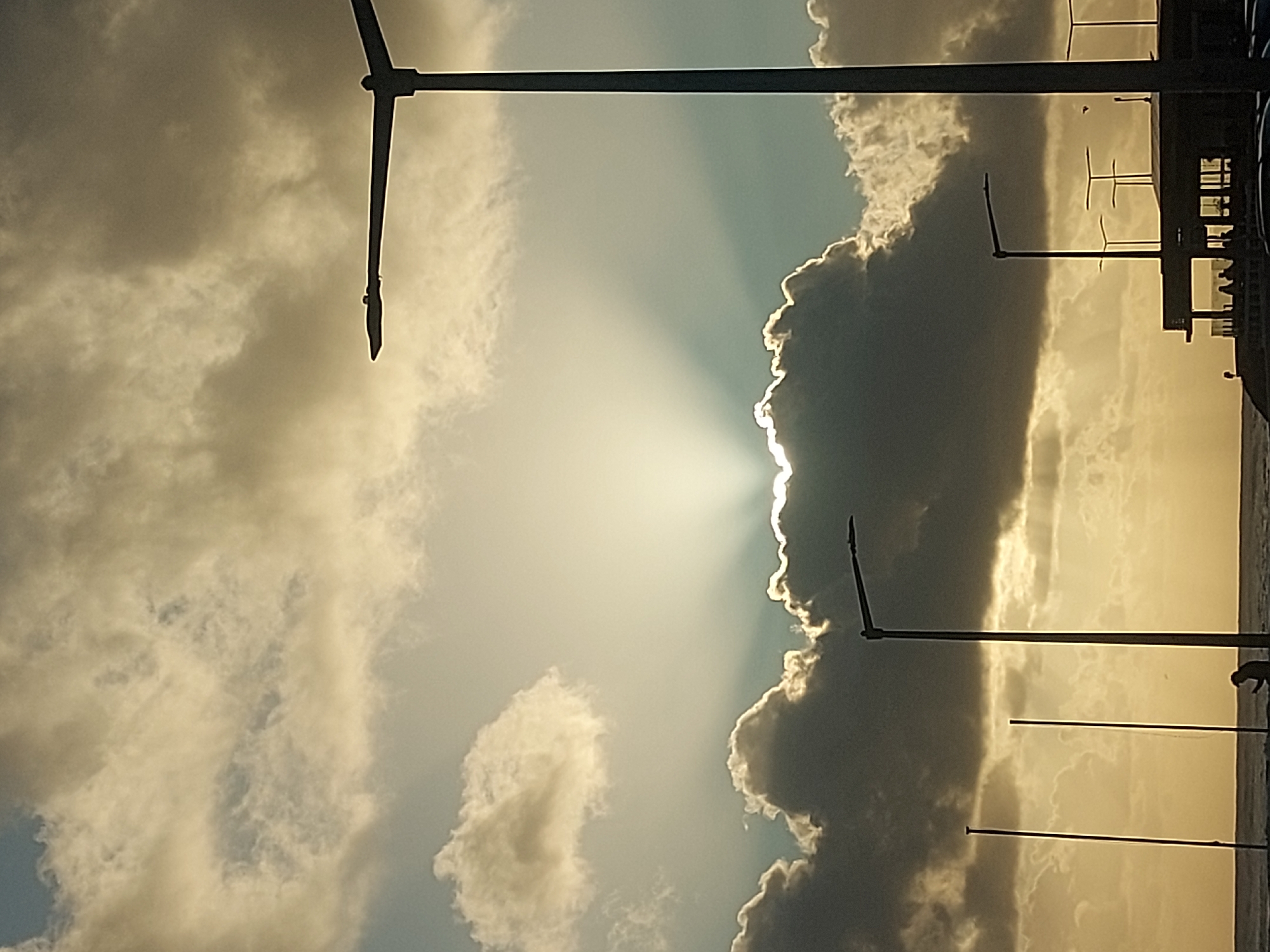 The image captures a sky view in Figueira da Foz, Portugal, dominated by heavy cloud cover. The sun's rays pierce through a gap in the clouds. Multiple streetlights are visible, their poles running vertically through the frame. On the right side, a building structure is discernible. A person is also visible towards the lower right corner of the image. The overall tone is subdued, with the sun providing a focal point.