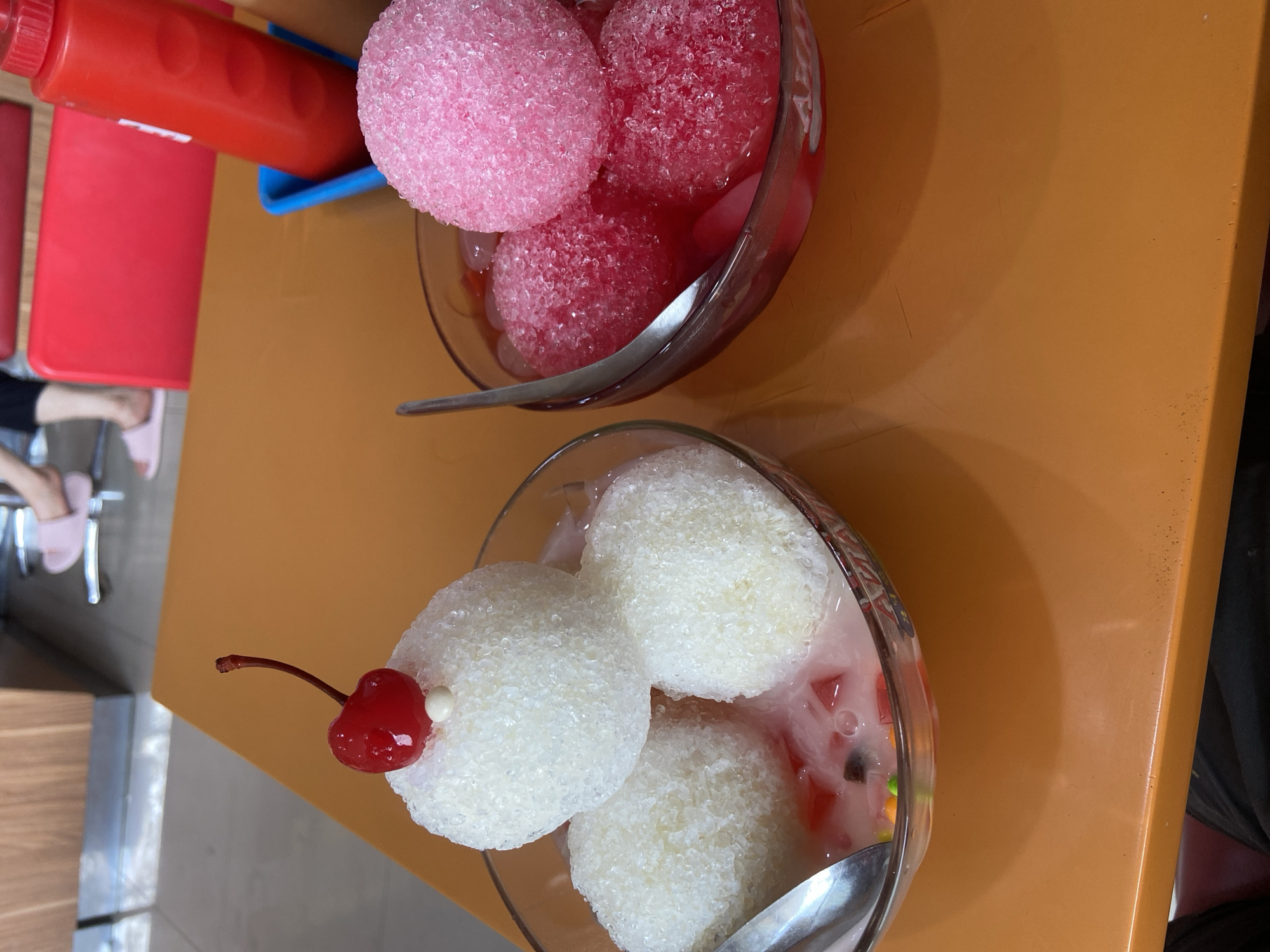 Two bowls of shaved ice desserts sit on an orange table in a restaurant setting, likely in Kudus, Indonesia. The bowl on the left contains three white scoops of shaved ice, topped with a maraschino cherry and a small white pearl. The ice is nestled in a milky liquid with colorful jelly pieces. The bowl on the right holds three pink scoops of shaved ice in a red, syrupy liquid. A red ketchup dispenser is visible in the background, along with part of a red chair and the lower legs and feet of a person wearing pink slippers. The overall environment suggests a casual dining establishment.