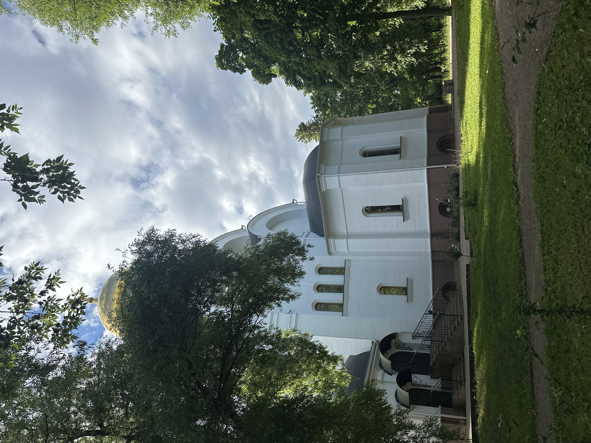 This is a vertically oriented, eye-level, outdoor photograph of a white Orthodox church with a golden dome. The church is partially obscured by lush green trees in the foreground, with their leaves creating a dappled effect on the building and the surrounding grass. Sunlight filters through the branches, casting shadows. The sky above is a mix of white and gray clouds.
The church itself has arched windows and a prominent golden dome visible above the trees. The building appears to be made of white stucco. There is a set of stairs leading up to an entrance on the right side of the building, with a black metal railing. The surrounding area is manicured with green grass and a pathway.
The time of day appears to be late morning or early afternoon, given the angle of the sun and the shadows. The weather is partly cloudy. There are no people visible in the image. No text is visible. The location context provided is Kotelniki, Russia, which aligns with the architectural style of the church. The overall mood is serene and tranquil, emphasizing the beauty of the church and its natural surroundings.