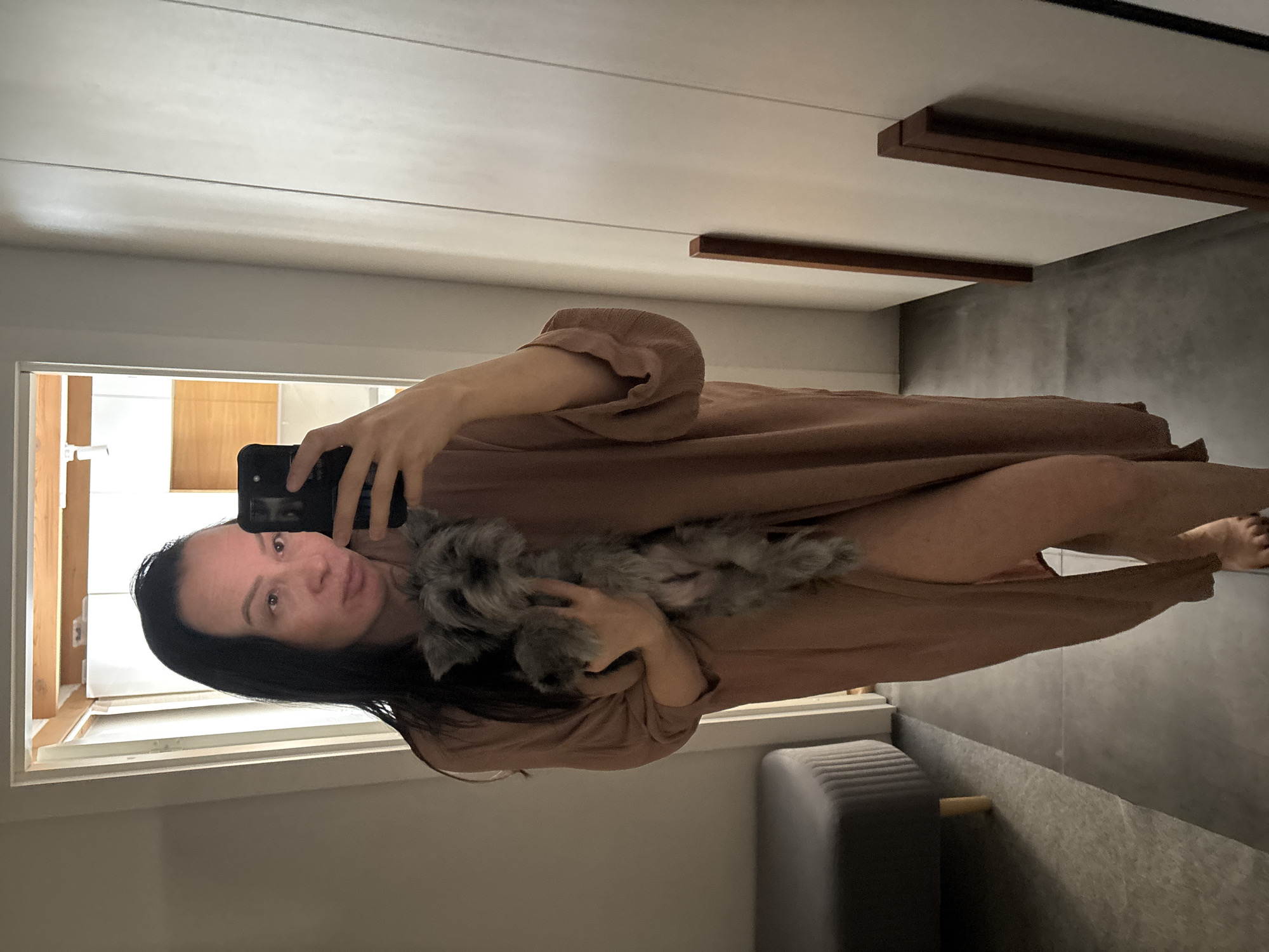 A woman with dark hair is taking a selfie in a well-lit interior. She is holding a small, gray, furry dog in her arms. She is wearing a brown robe or dress. The background includes a door frame with a view into another room with wood paneling, a gray stool, a gray wall, and ceiling trim. There is not sufficient information to determine if the location is in the city of Chaikovsky, Russia.