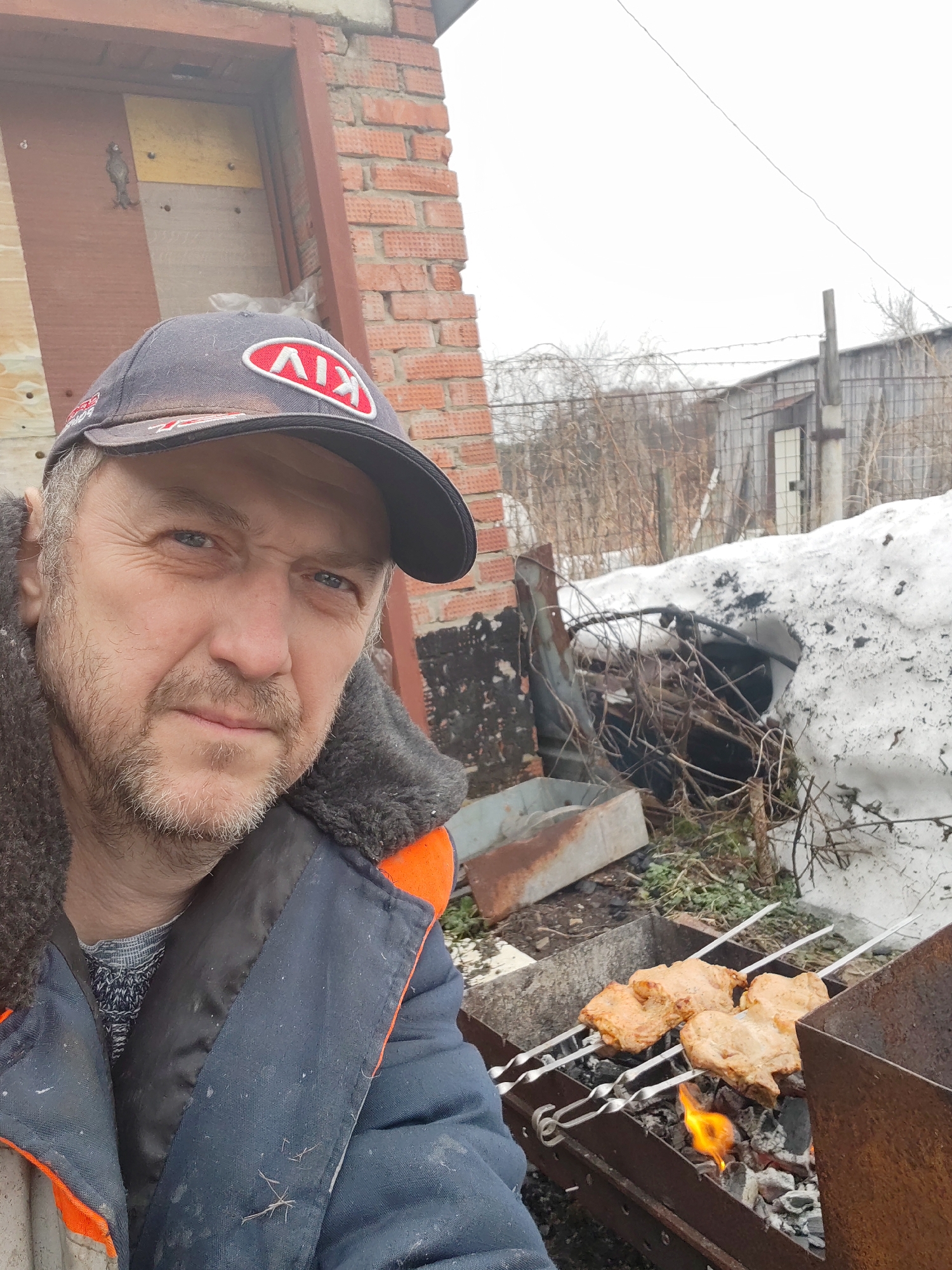 The image is a selfie of a middle-aged man wearing a dark blue jacket with orange trim, a cap with the "KIV" logo, and a grey patterned sweater. The background reveals a backyard scene with a barbecue grill.
On the grill are several metal skewers holding pieces of what appears to be marinated meat, cooking over glowing embers and small flames. The surrounding area includes a pile of snow, dry vegetation, a partially visible building with a brick wall, and a metal fence with a barbed wire.
The photograph was taken outdoors in the city of Chaikovsky, Russia.