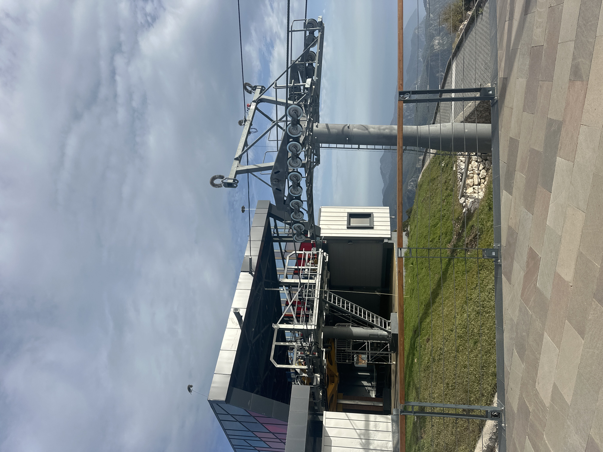 The image depicts a mountain cable car station situated near Fethiye, Turkey. The station features a modern architectural design with glass and metal paneling. A prominent cable car tower supports a complex system of cables and pulleys for transporting passengers. A grassy slope and a stone-paved area surround the station, with a metal fence delineating the boundaries. Distant mountains are visible in the background under a cloudy sky.