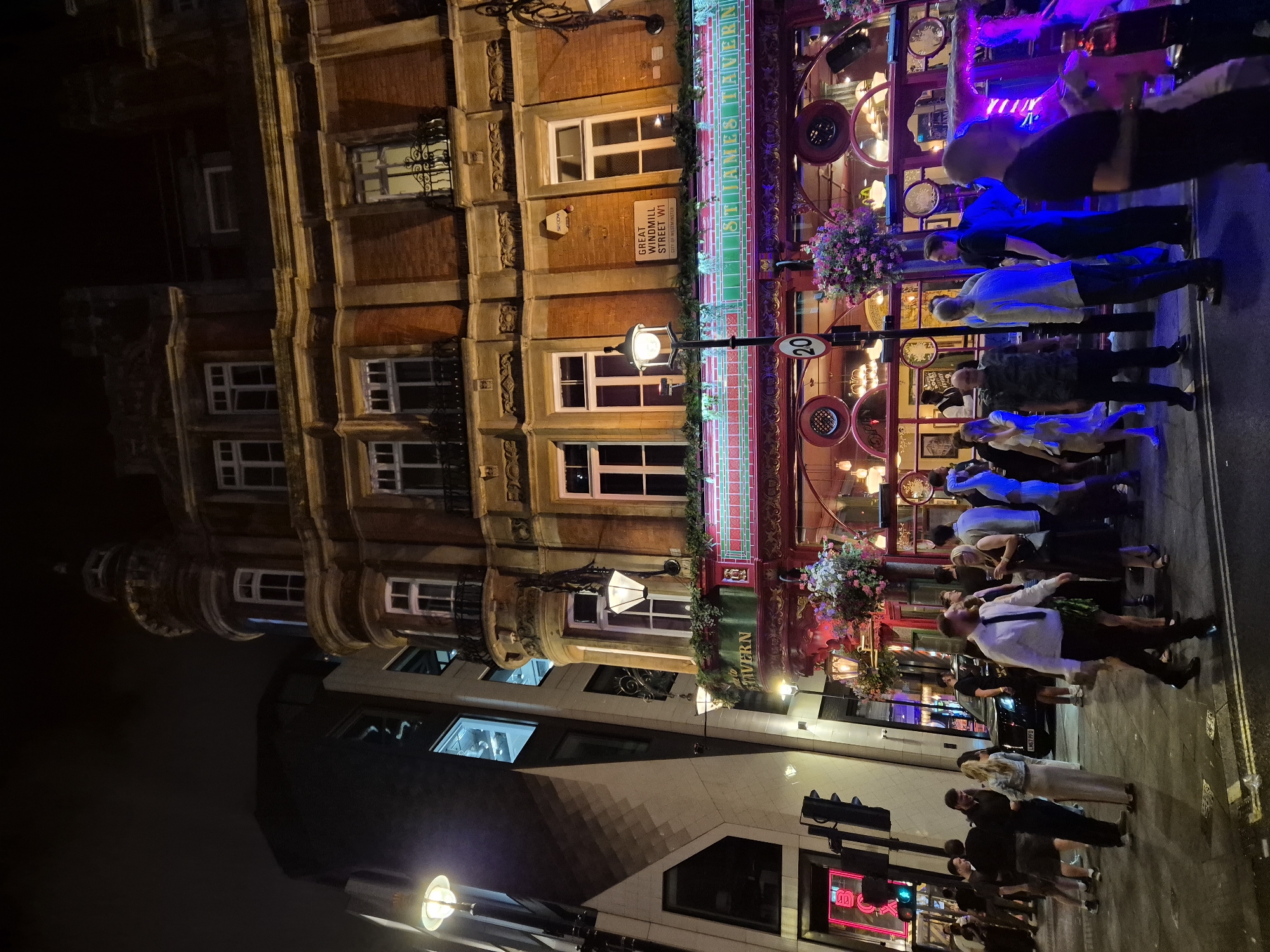 A street-level, medium shot captures the exterior of "St. James Tavern" in the city of London, United Kingdom, at night. The tavern, a brick building with ornate architectural details, is illuminated by neon lights and streetlamps. A diverse group of people are seen walking and standing on the pavement in front of the establishment. The atmosphere suggests a lively evening scene, with a mix of casual and dressed-up individuals. The street is paved, and traffic lights are visible in the background, indicating a public thoroughfare. The image is characterized by strong contrasts between light and shadow, typical of a nighttime urban environment.