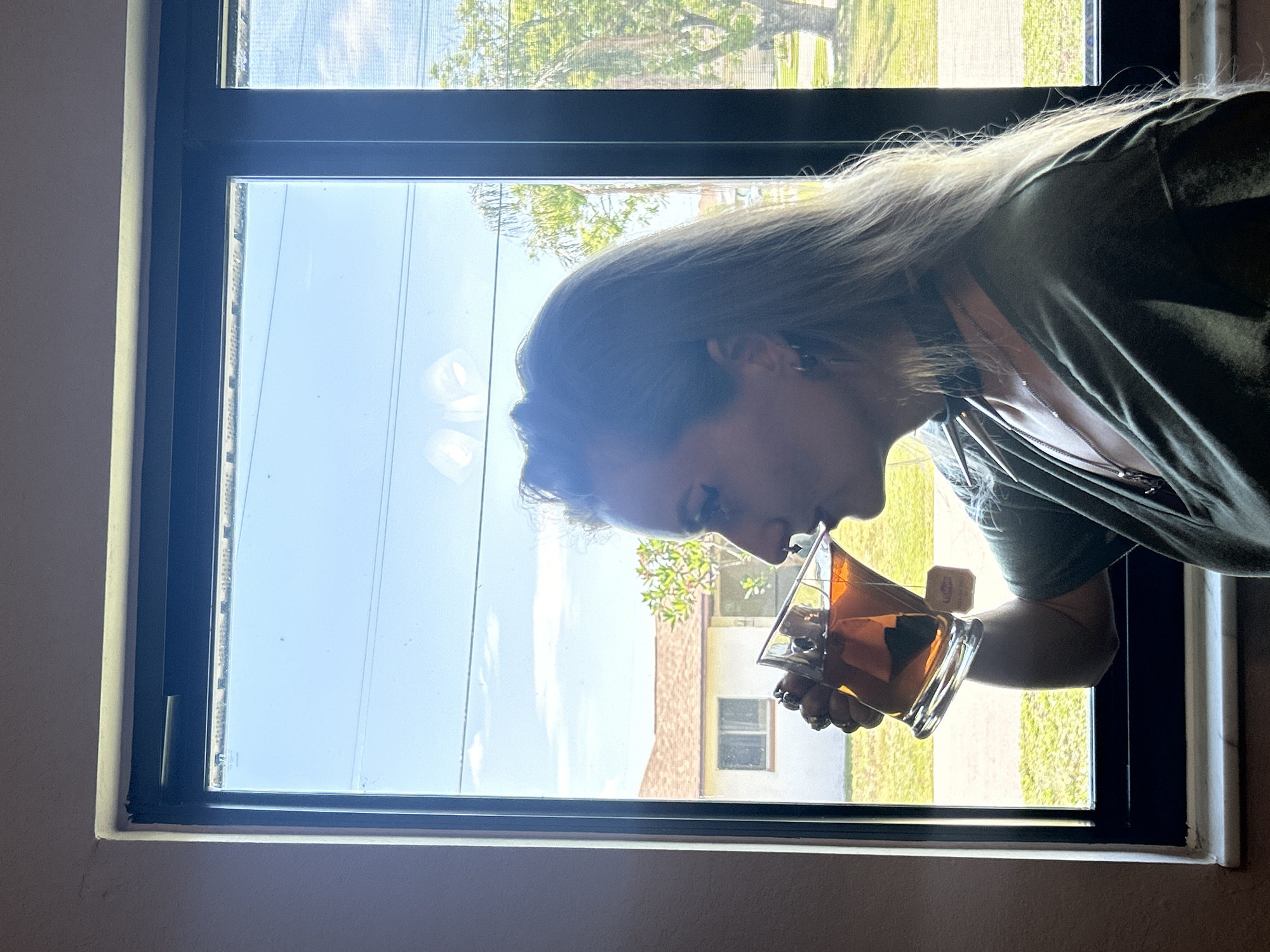 A woman is pictured from the chest up, from the left side, drinking from a clear glass filled with amber liquid and a tea bag. She has long blonde hair, is wearing a green top, a black choker necklace, and a silver necklace with a pendant. Her eyes are closed as she drinks. She is standing in front of a large window with a dark frame, looking out at a sunny day in Cape Coral, Florida. Through the window, a white house with a red-tiled roof, green grass, trees, and a bright blue sky with some clouds are visible.