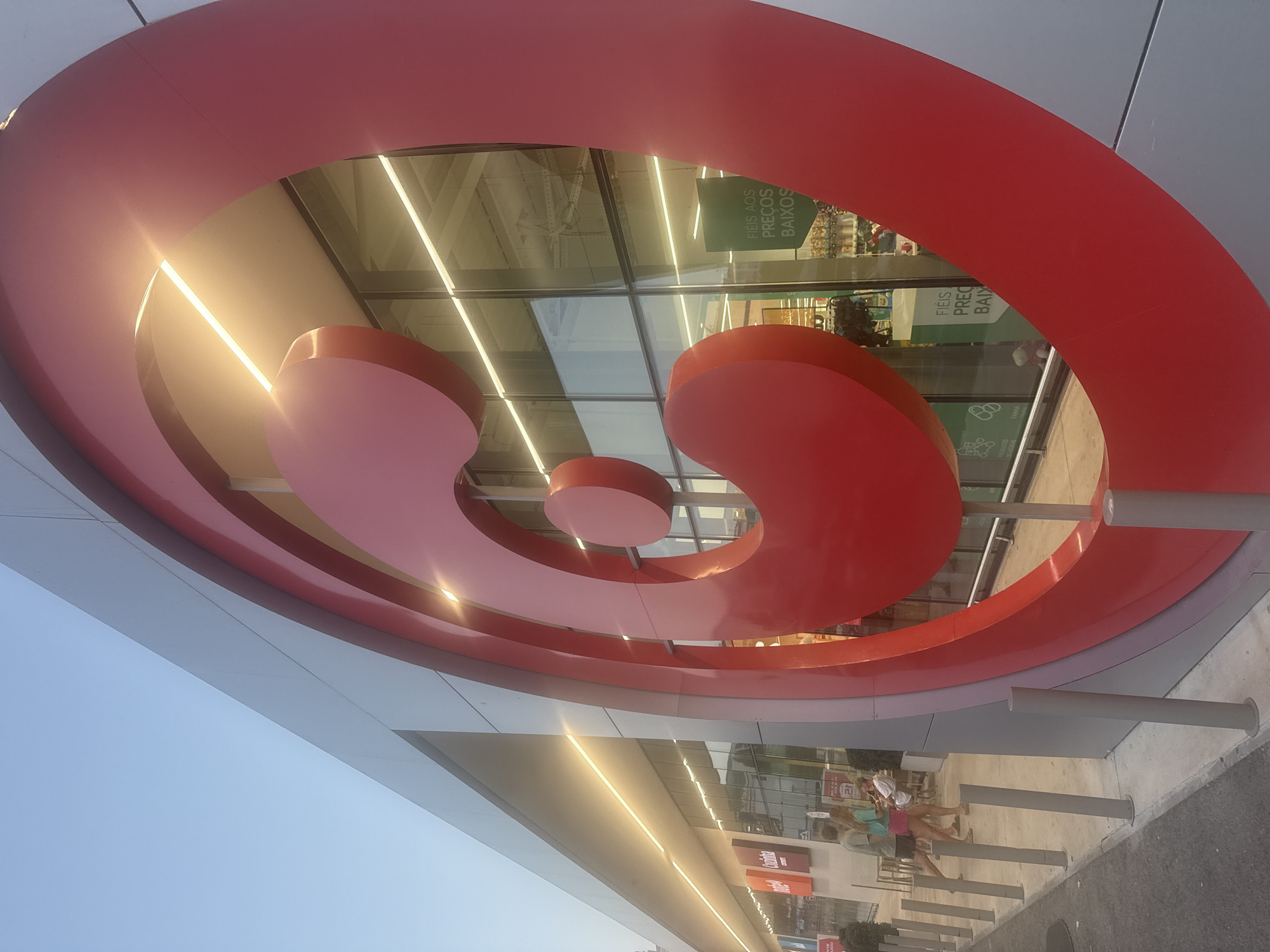 The image shows the exterior of a Pingo Doce supermarket in Torres Vedras, Portugal, during the evening. A large, red, circular logo, characteristic of the supermarket chain, dominates the foreground, angled towards the viewer. The building's facade is silver-toned metal paneling, with large glass windows reflecting the interior lighting and some of the exterior environment. Inside the store, aisles of merchandise are visible. A green sign with white lettering that reads "FIÉIS AOS PREÇOS BAIXOS" (Loyal to low prices) is partially visible through the glass. To the left of the main logo, a portion of the building's entrance area is visible, with several orange rectangular signs displaying text in Portuguese, including "Cozinha". Outside the entrance, a few people are standing and walking. A sidewalk with bollards separates the building from a paved area, likely a parking lot or pedestrian walkway. The sky above is a pale blue, indicating twilight or early evening.