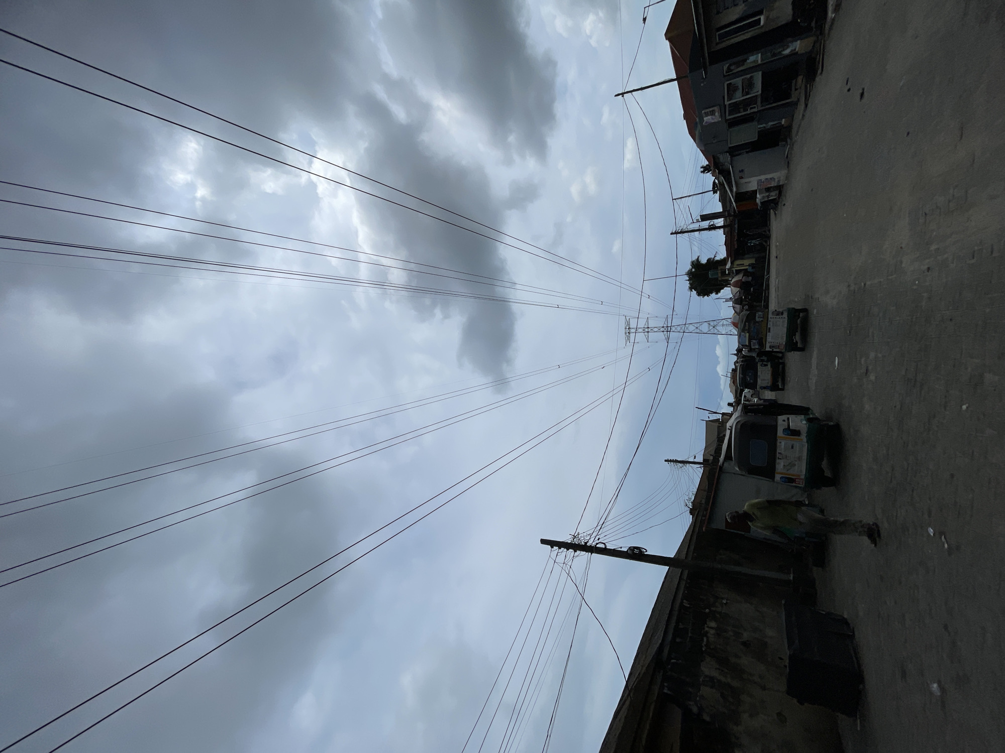The image depicts a street scene in Ado, Nigeria, characterized by a combination of infrastructure and everyday activity. Overhead, high-voltage power lines converge towards a tall transmission tower in the distance under a cloudy sky. Below, a road stretches between buildings, with evidence of pedestrian and vehicular traffic. An individual walks along the side of the road near a building. Several auto-rickshaws, commonly known as "Keke Napep," are parked along the roadside. Utility poles supporting secondary power lines are also visible along the street.