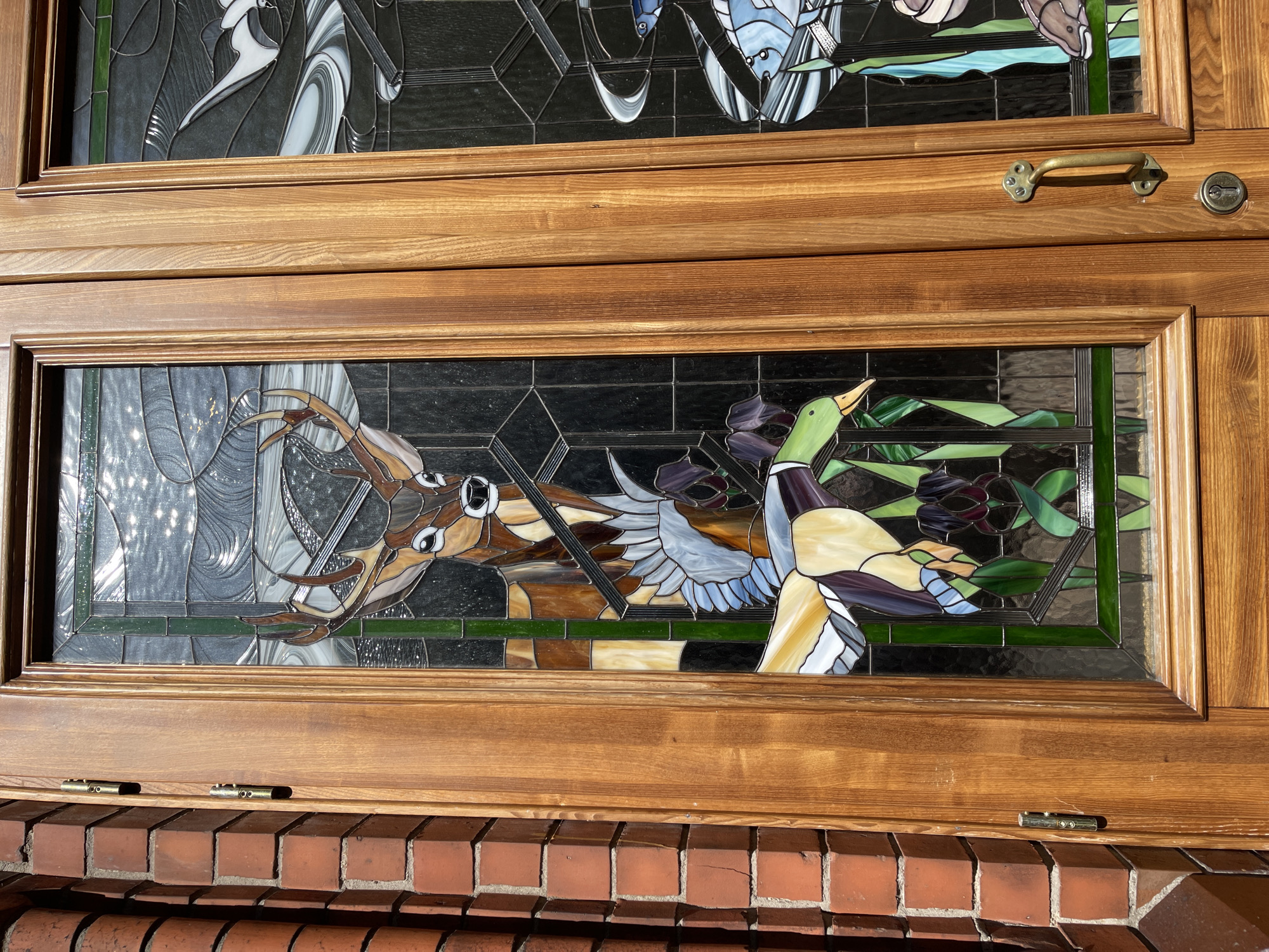 The image shows a close-up of a wooden double door with stained glass panels. The left panel features a stained glass depiction of a deer with antlers, a bird with blue and white feathers, and a duck with green and yellow plumage in front of dark green reeds and purple flowers. The right panel, partially visible, shows stained glass images of fish and birds in a watery environment. The doors are set against a brick wall. A brass door handle and a keyhole are visible on the right door. The scene is outdoors, with sunlight glinting off the stained glass. This is likely a residential or public building entrance in Zelenogradsk, Russia.